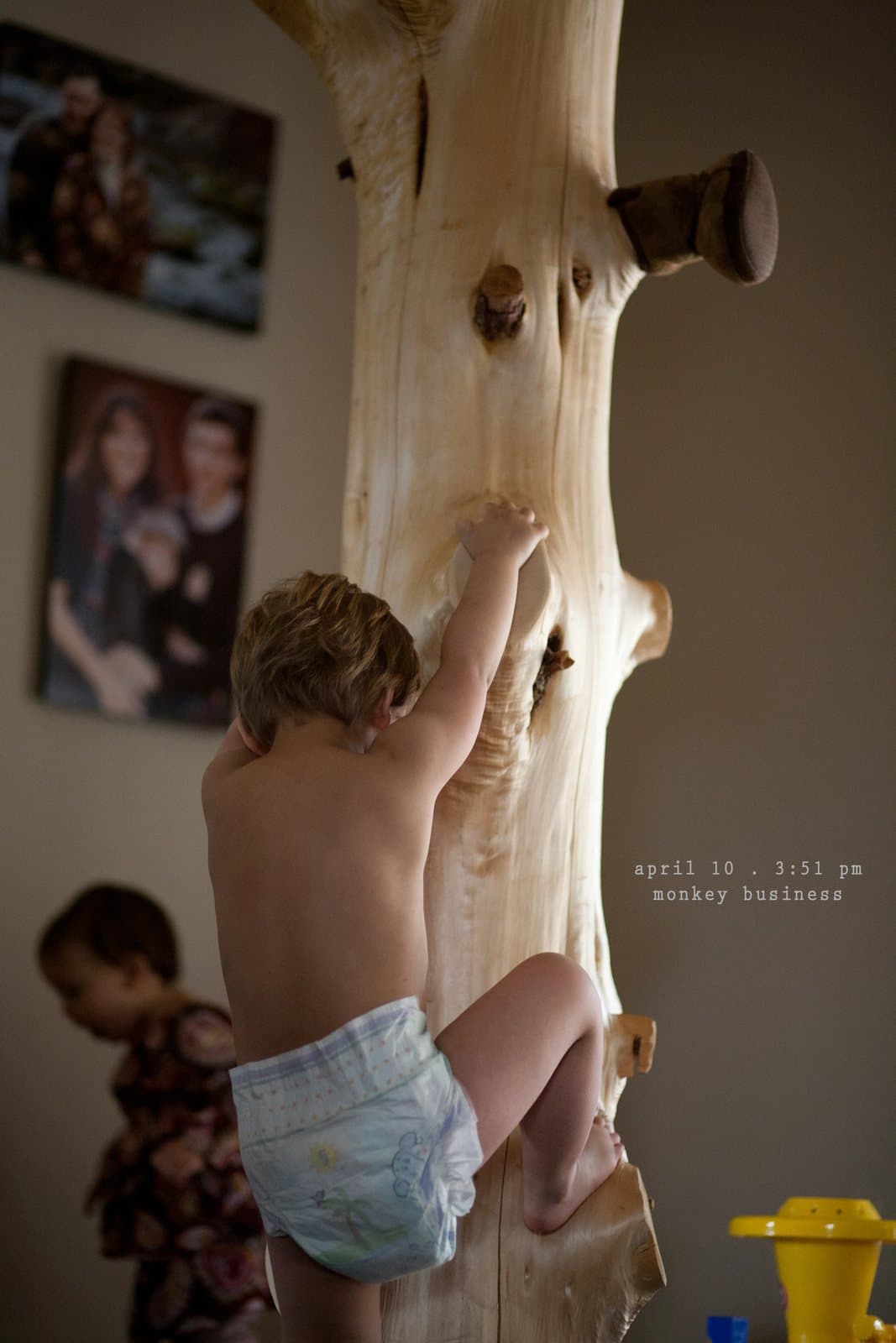 A toddler in a diaper climbs a carved log pole in a living room, captioned monkey business.