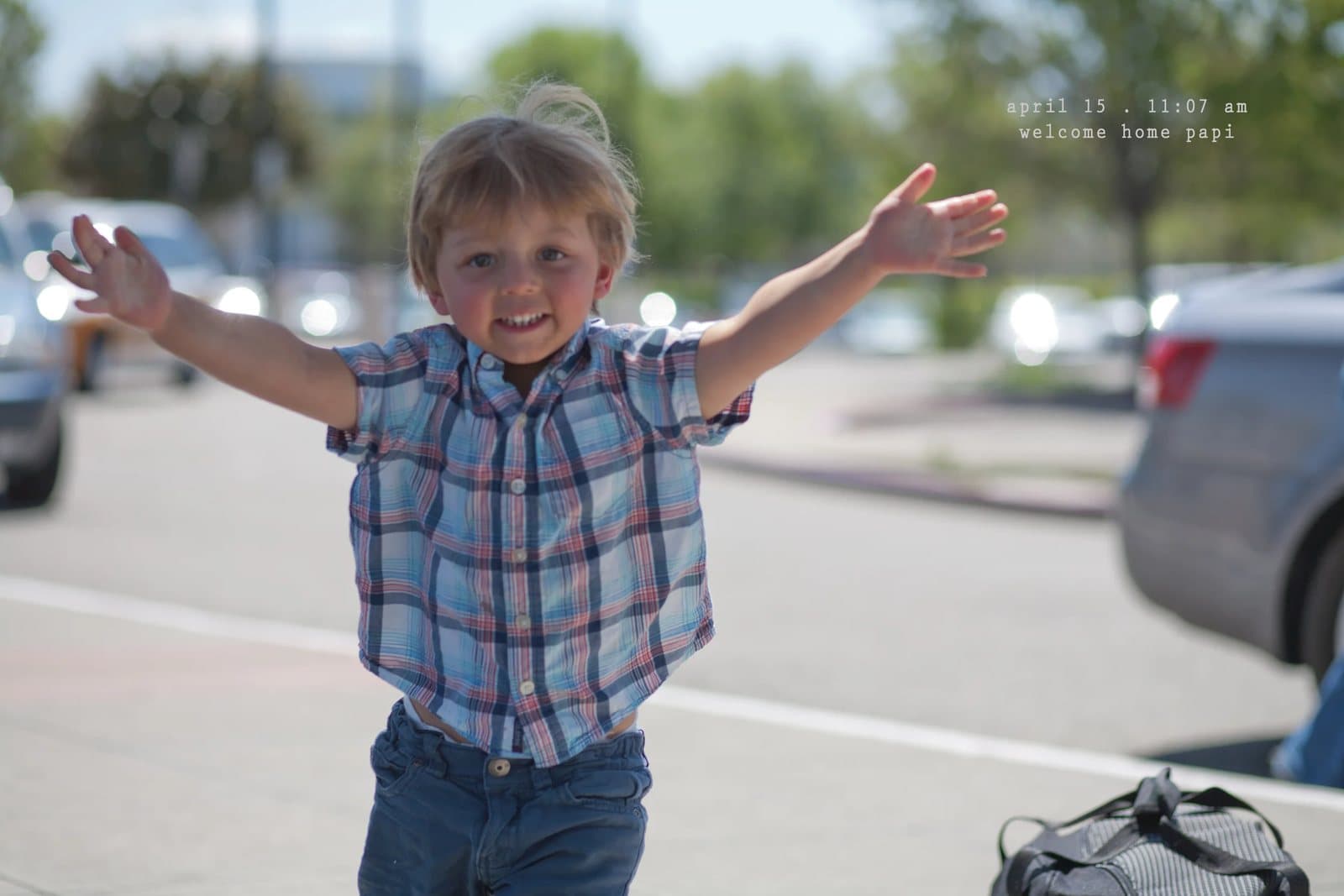 A smiling toddler throws his arms wide in a parking lot, captioned welcome home papi.