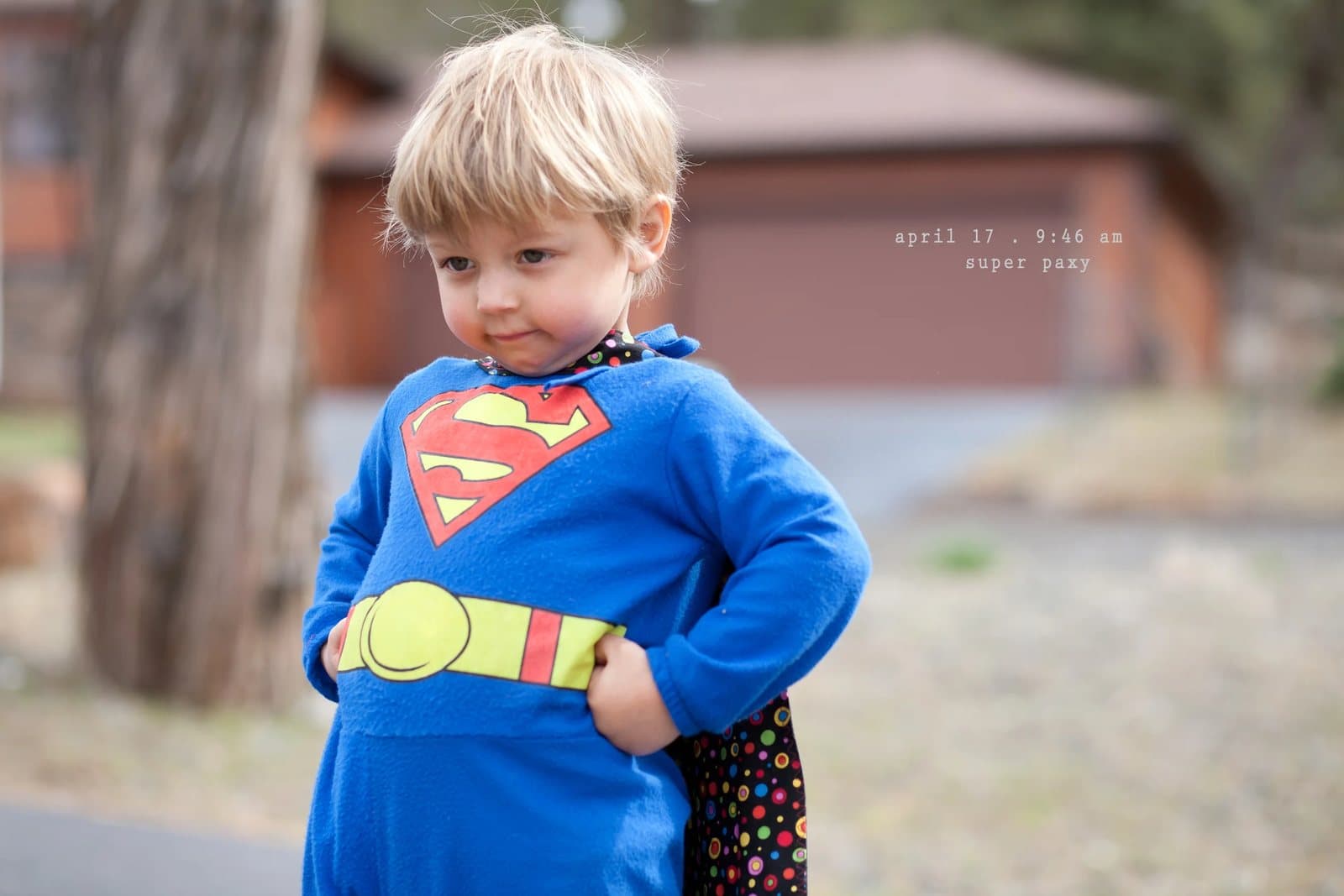 A blond toddler in a Superman costume stands with hands on hips, captioned super papi.