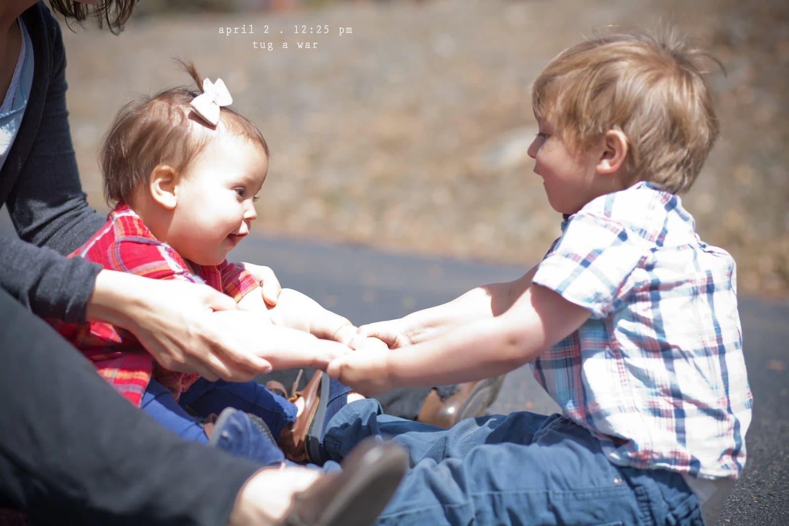 A baby girl with a bow and a toddler boy hold hands in a tug of war.