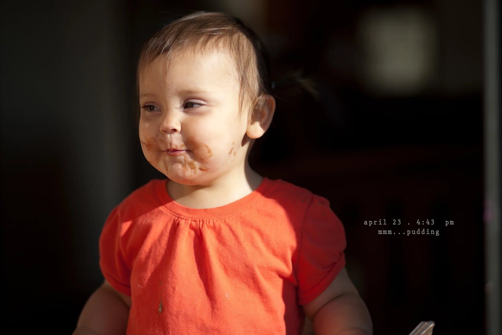A baby in an orange shirt with a pudding-smeared face, captioned mmm pudding.