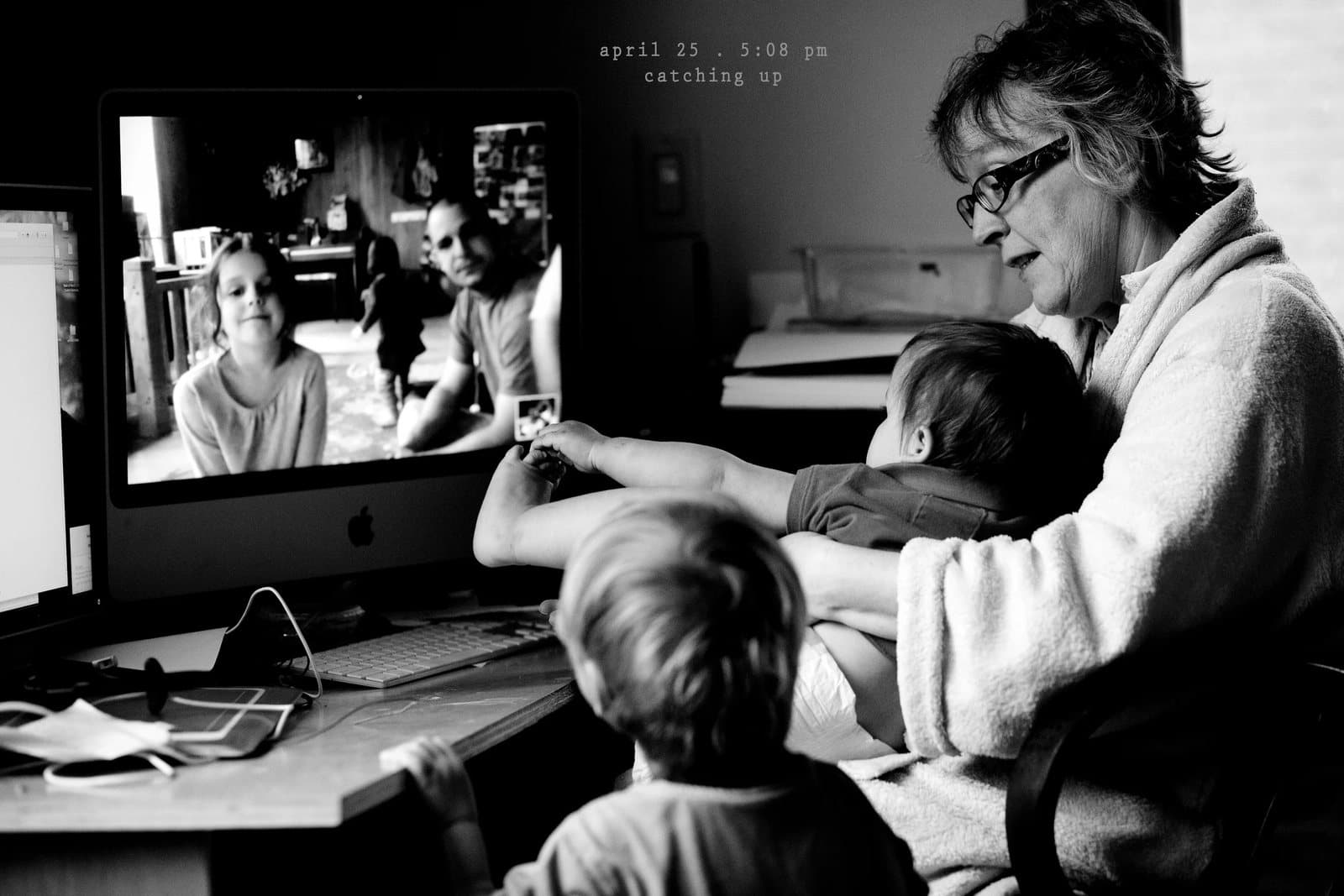 A grandmother holds two children at a computer showing a video call, captioned catching up.
