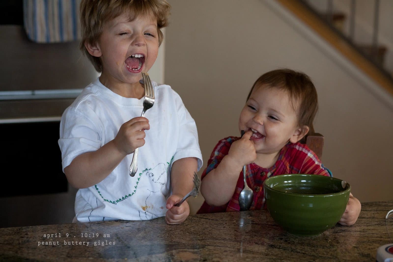 A boy and a baby laugh while eating peanut butter at a counter, captioned peanut buttery giggles.
