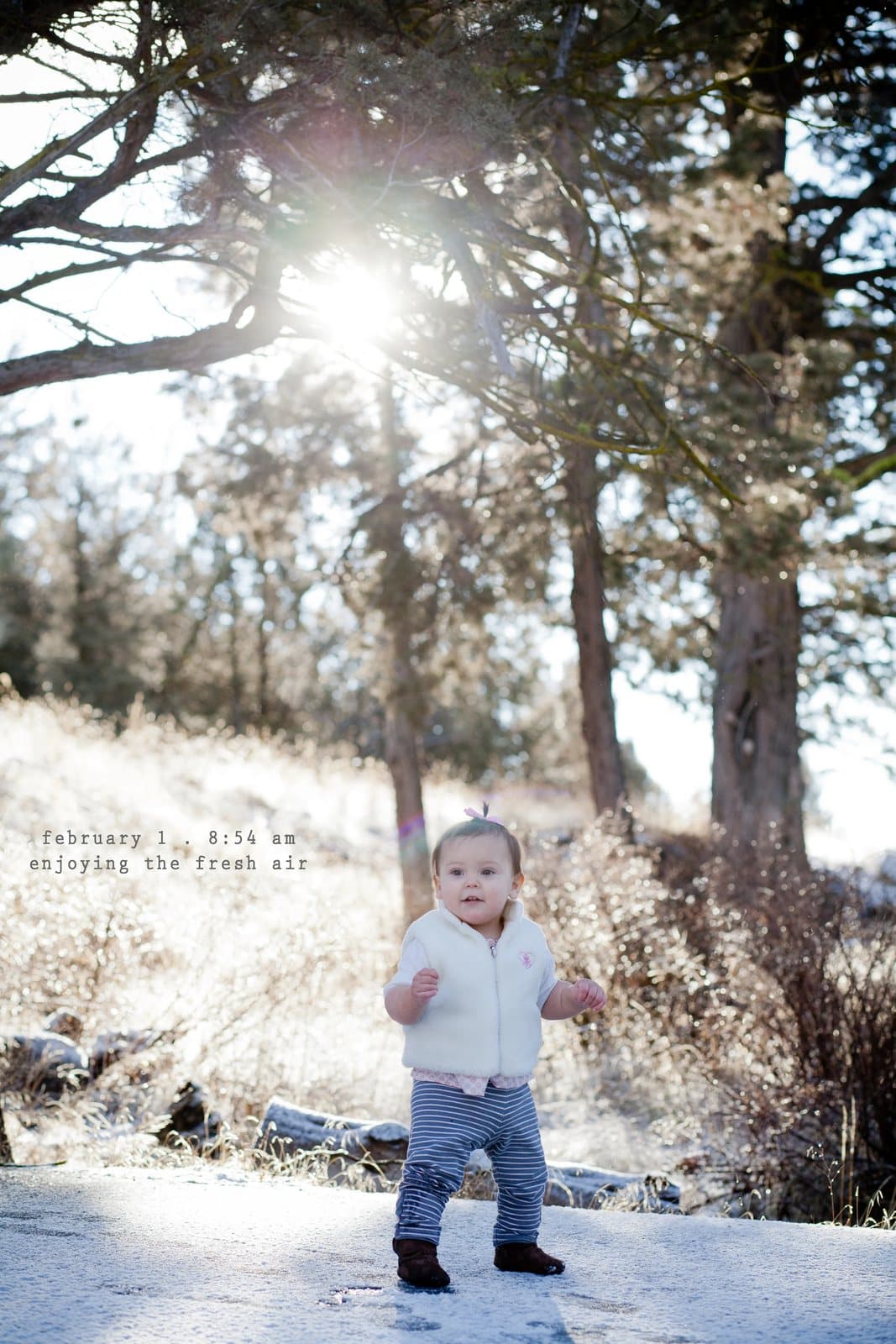 A baby in a white vest stands on a sunlit path in the snow, captioned enjoying the fresh air.