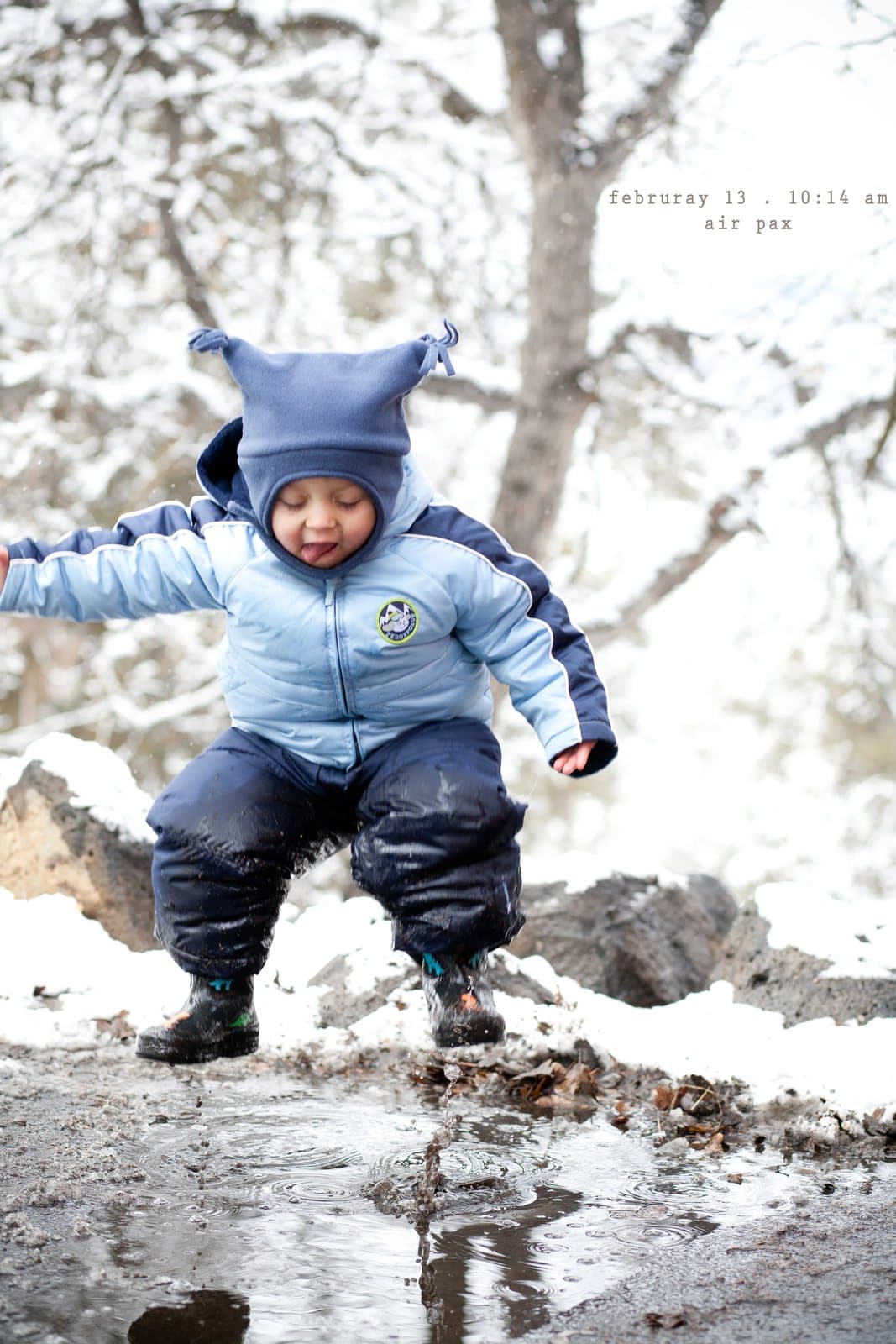A toddler named Pax in a blue snowsuit jumps into a puddle, captioned air pax.