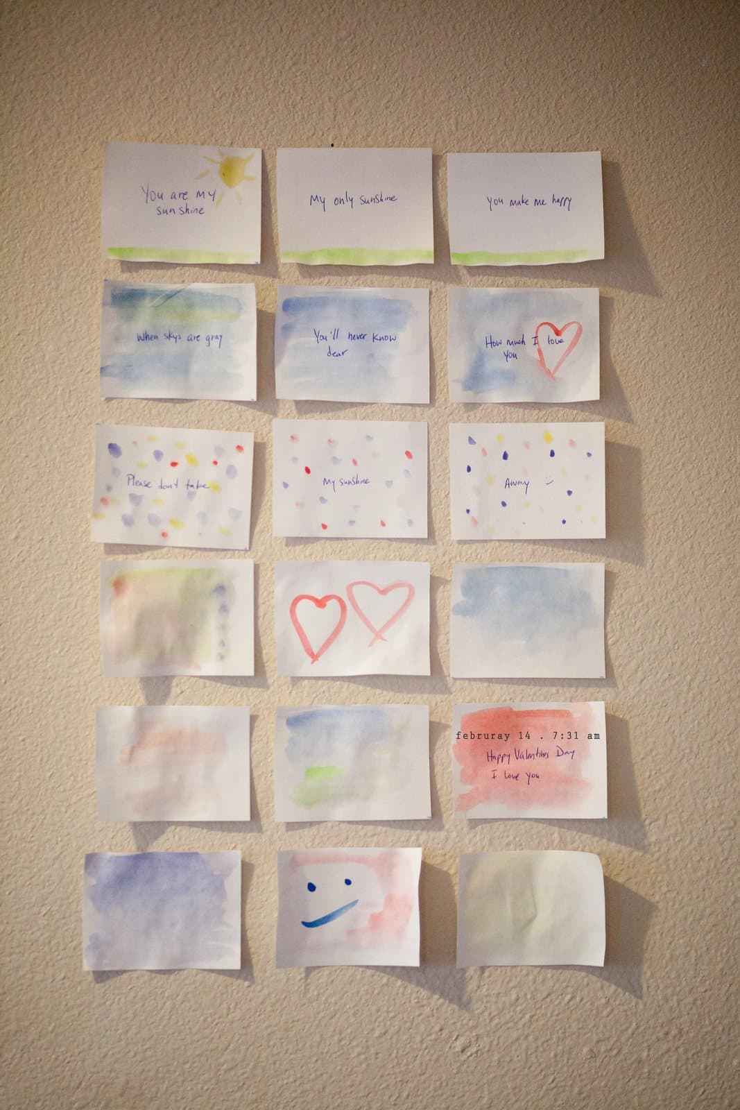 A grid of small watercolor love notes taped to a wall, including Happy Valentine's Day, I love you.