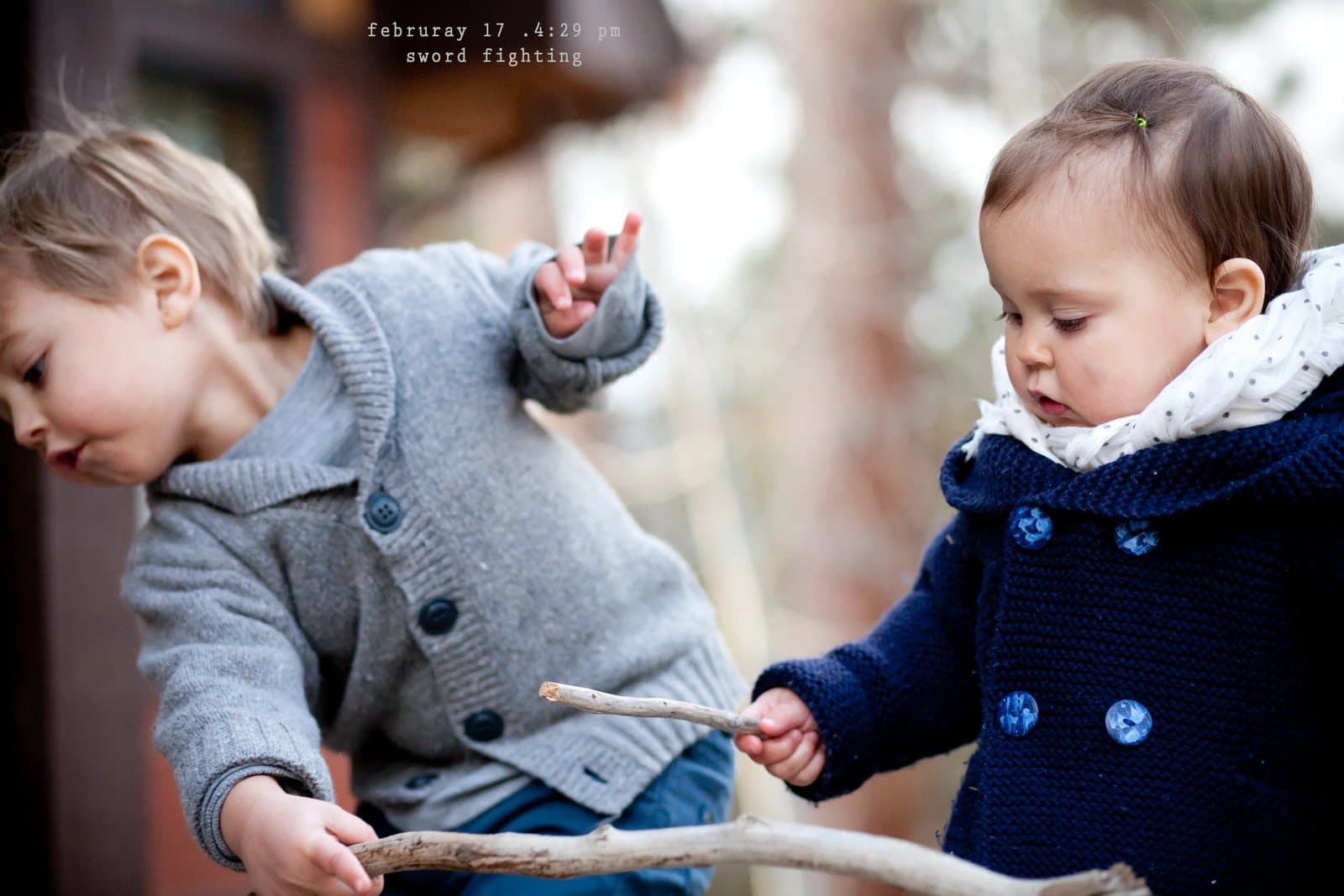 A toddler boy and a baby girl in winter coats play with sticks, captioned sword fighting.