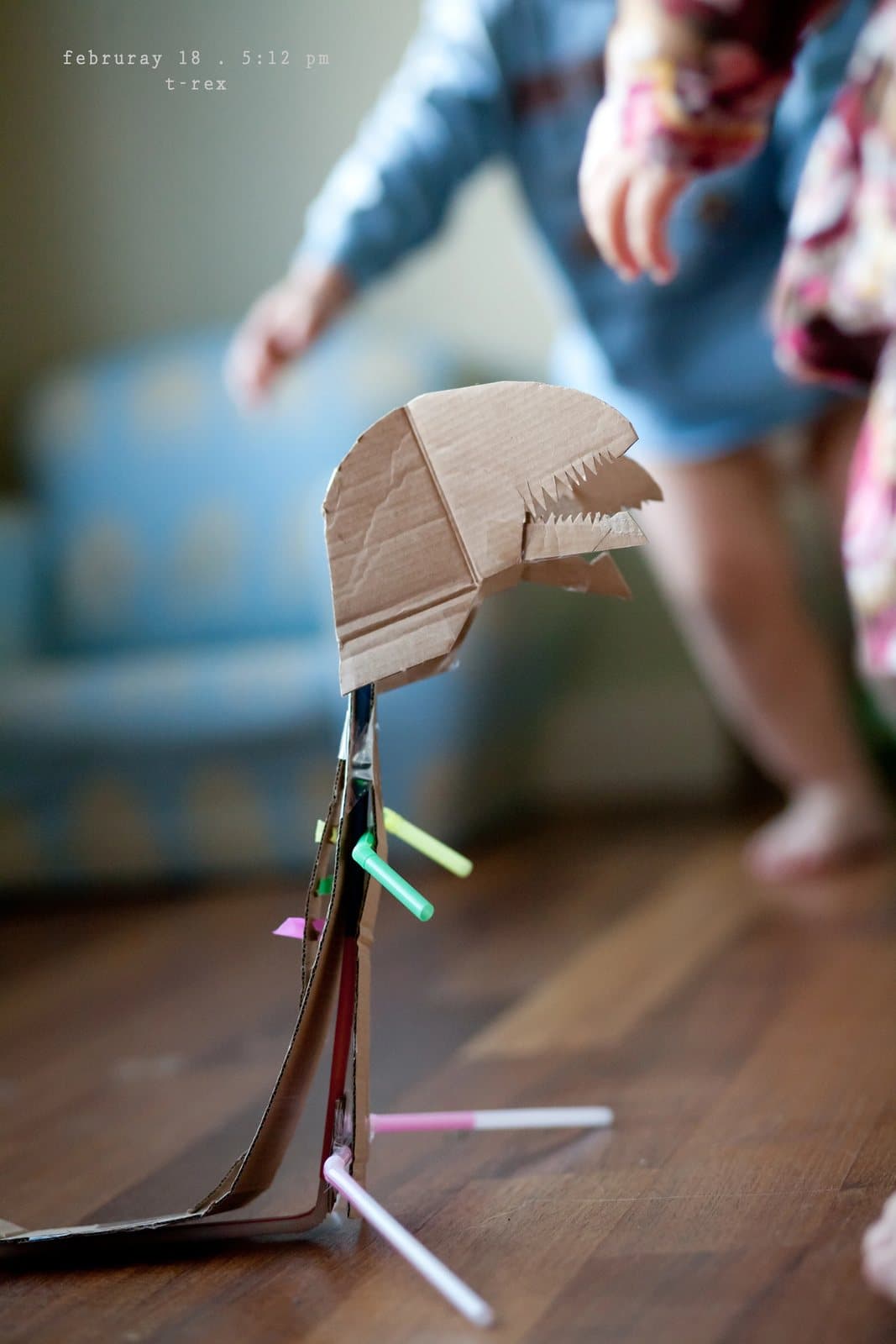 A cardboard T-rex head on a stick with colored straws stands on a wood floor.
