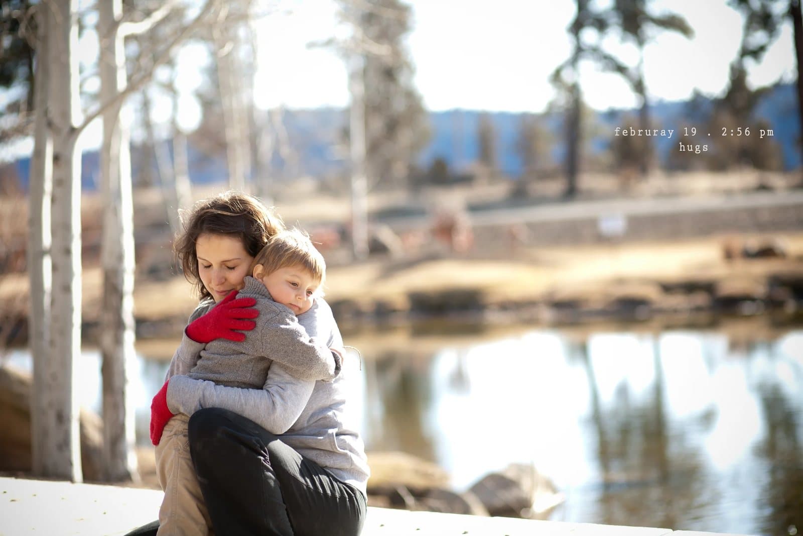 A woman hugs a toddler in a gray sweater beside a pond, captioned hugs.