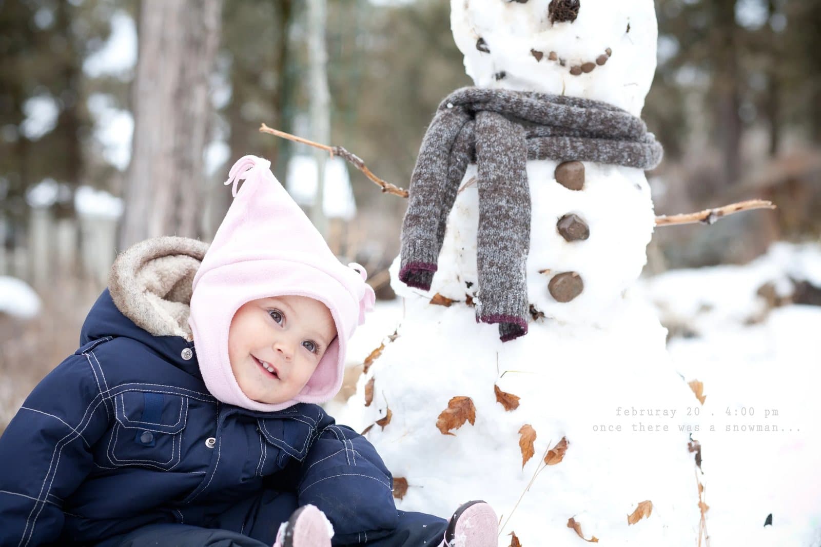 A smiling baby in a pink hat sits beside a snowman wearing a scarf.