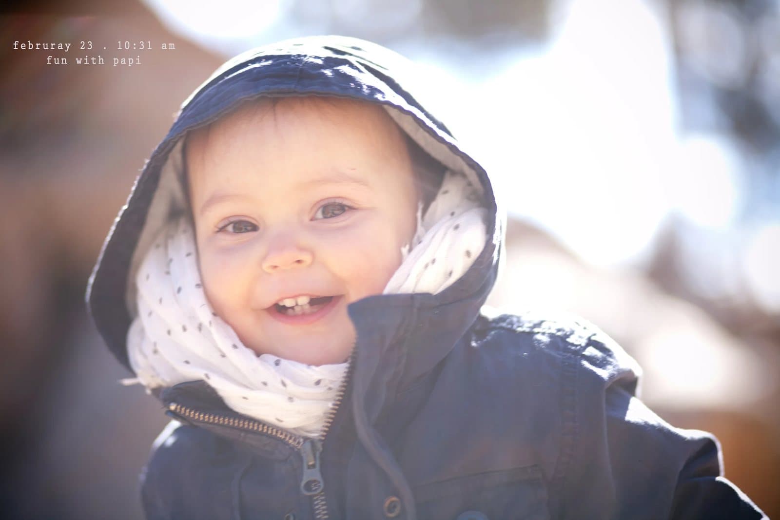 A grinning baby in a hooded coat and scarf in bright sun, captioned fun with papi.