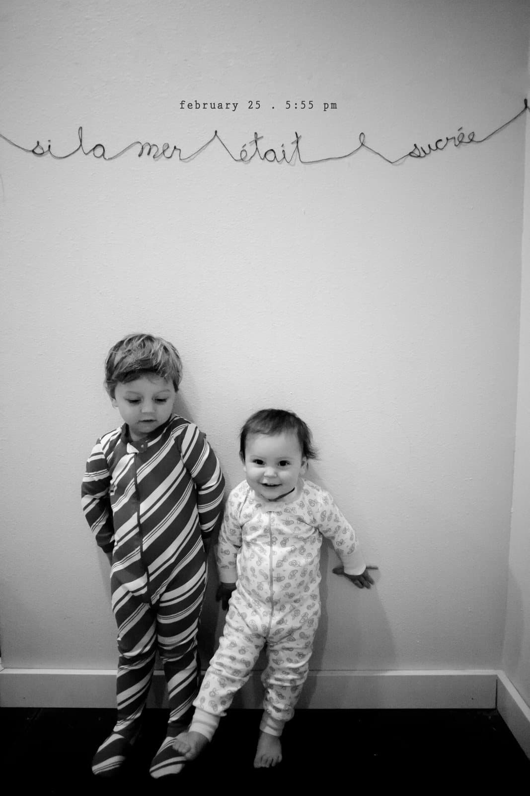 Two children in pajamas stand under the wall lettering si la mer etait sucree.