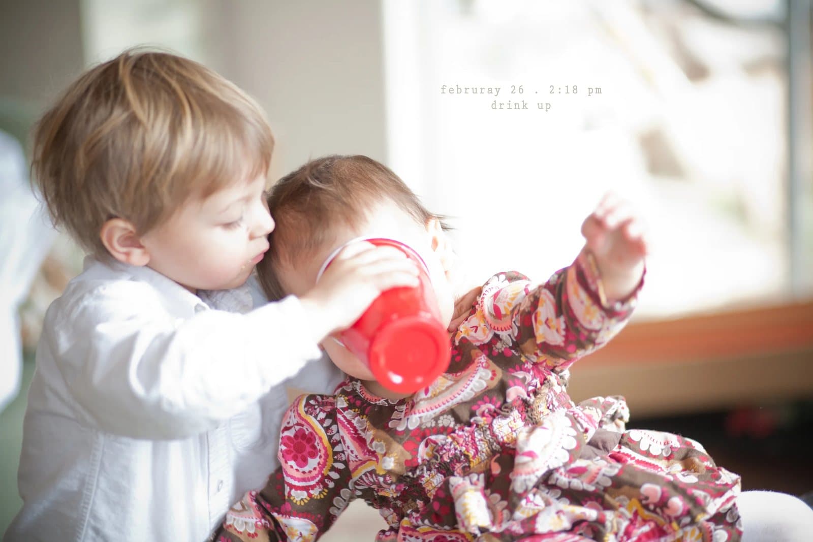 A toddler helps a baby drink from a red cup, captioned drink up.