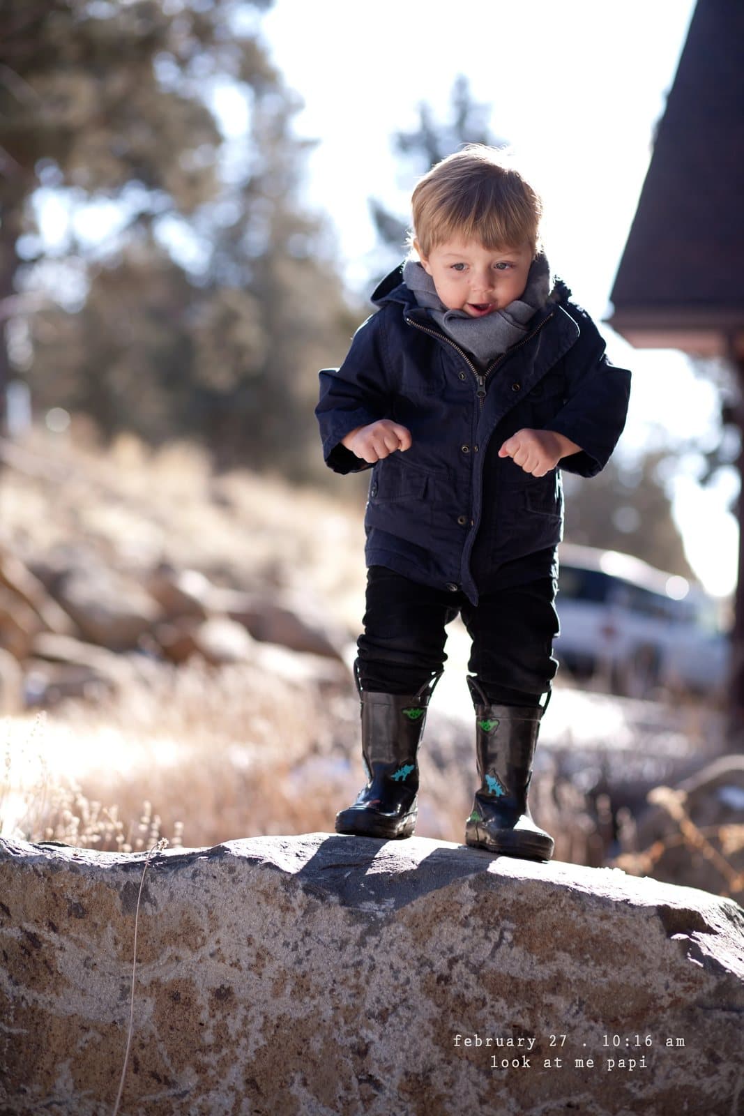 A toddler in rain boots stands on a rock, captioned look at me papi.