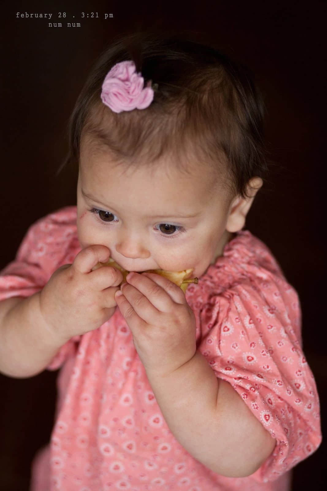 A baby with a pink flower clip eats with both hands, captioned num num.