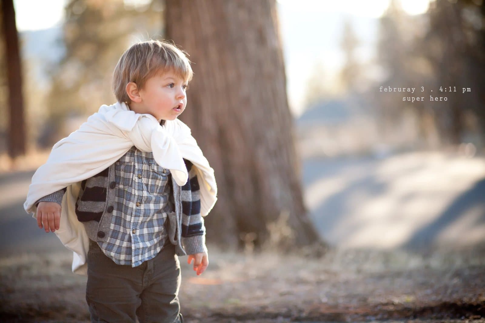 A toddler wearing a white cape walks among pine trees, captioned super hero.