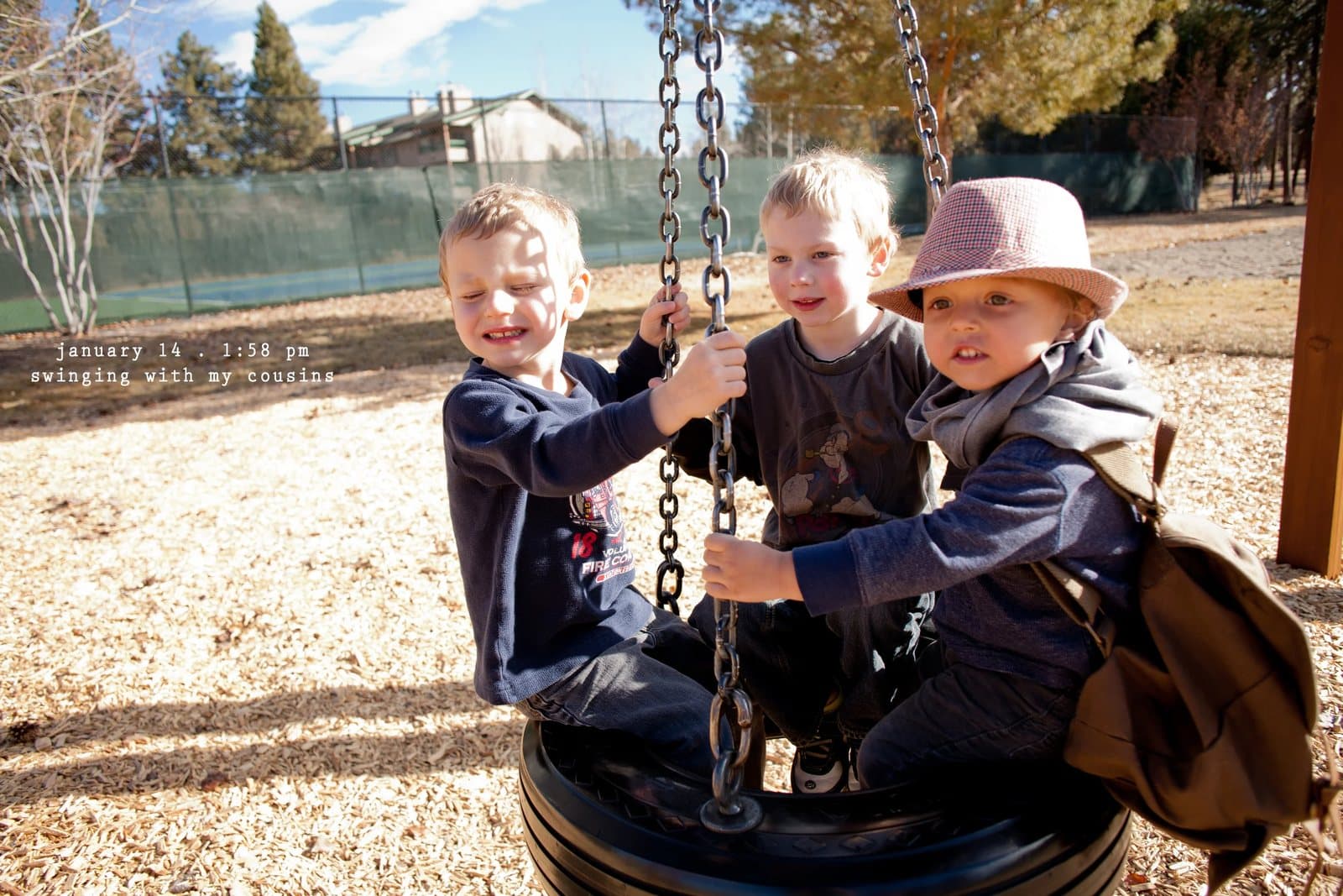 Three young boys ride a tire swing at a playground, captioned swinging with my cousins.