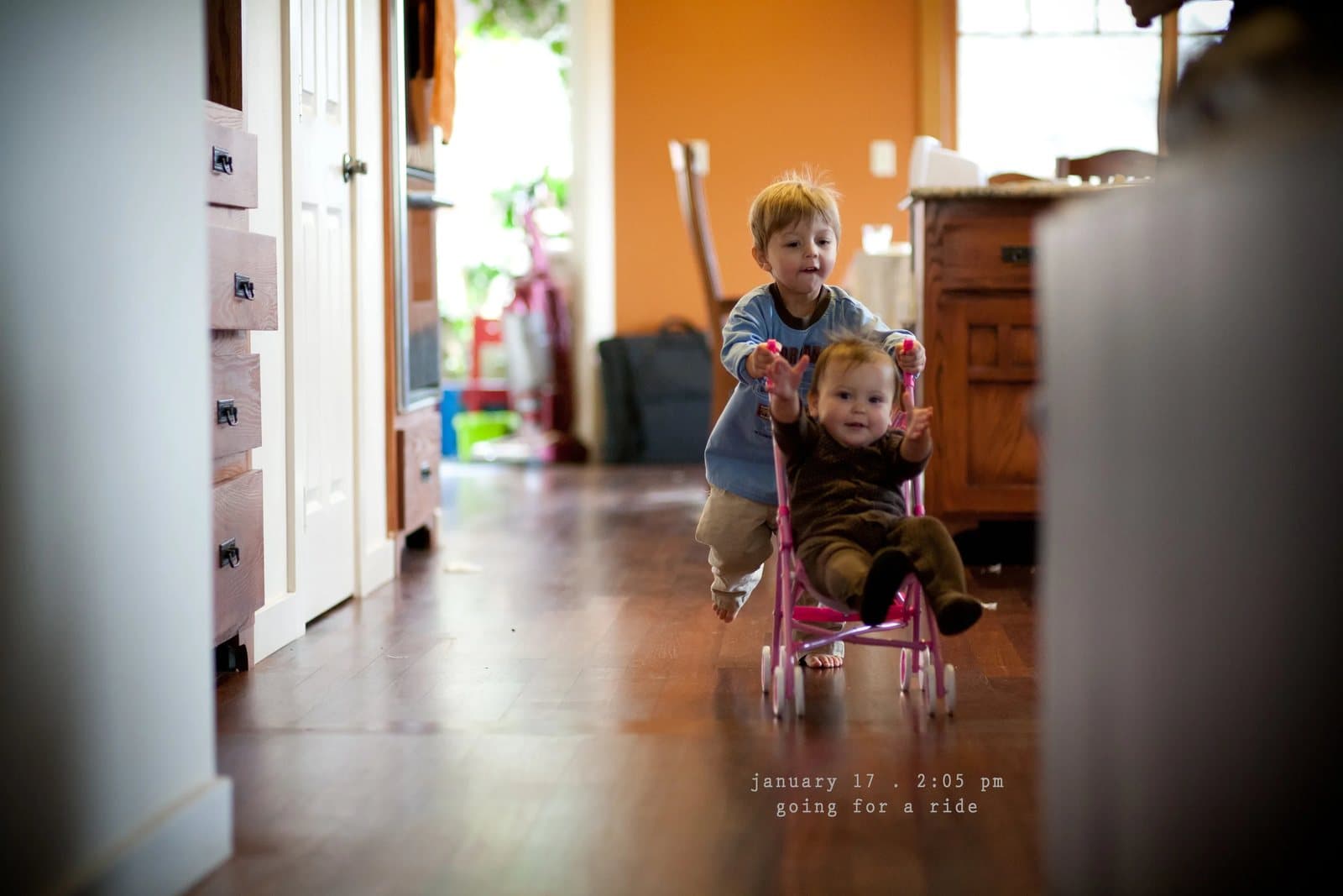 A toddler pushes a baby in a pink toy stroller down a hallway, captioned going for a ride.