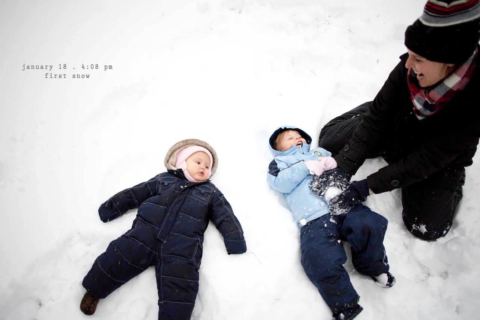A woman plays with two babies in snowsuits lying in the snow, captioned first snow.
