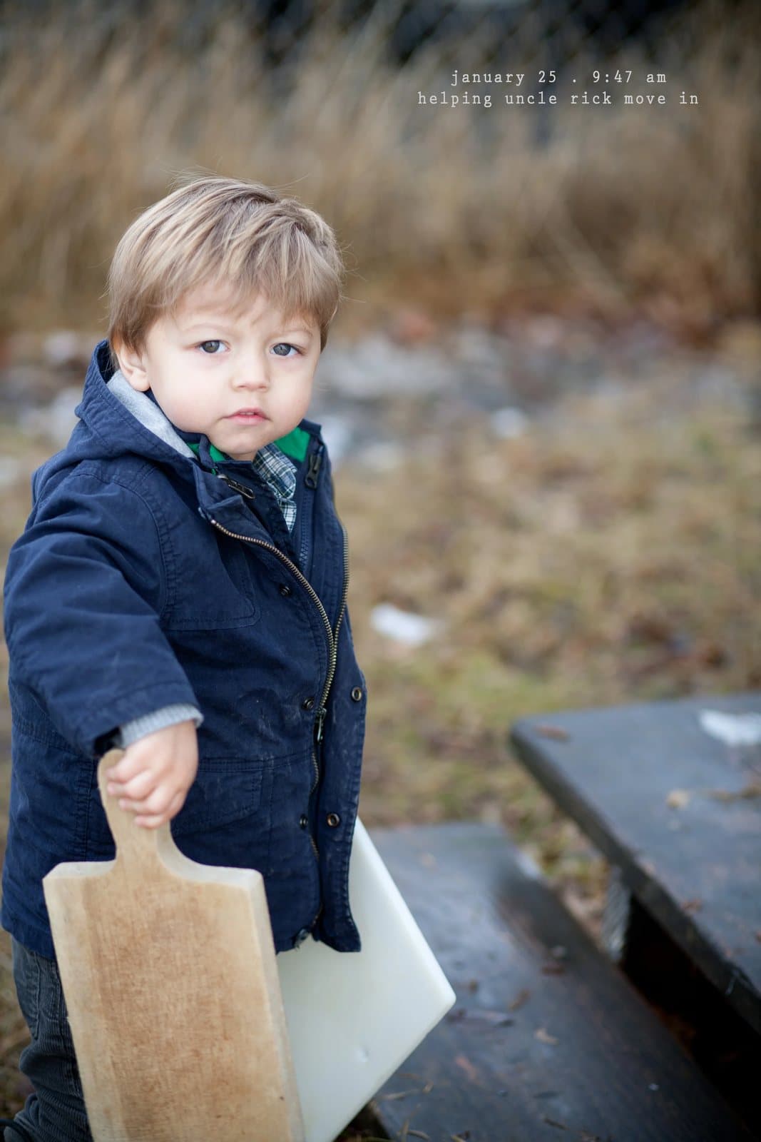A toddler in a navy coat carries a cutting board outdoors, captioned helping uncle rick move in.