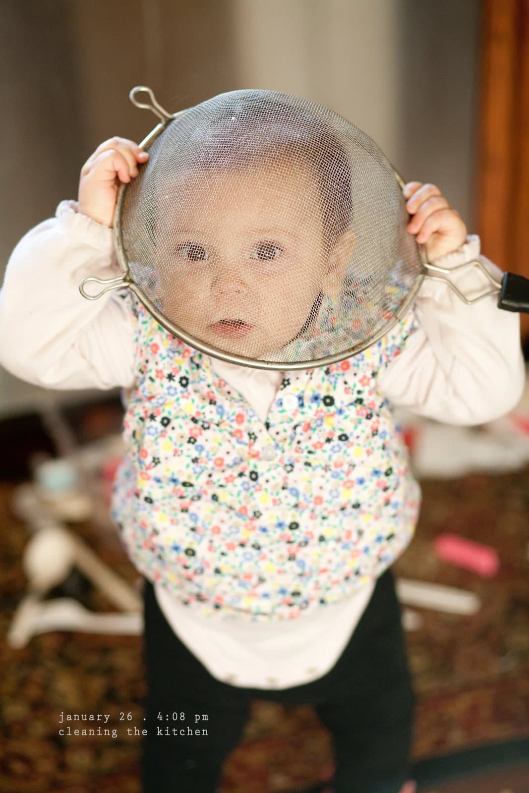 A baby holds a mesh strainer over her face like a mask, captioned cleaning the kitchen.