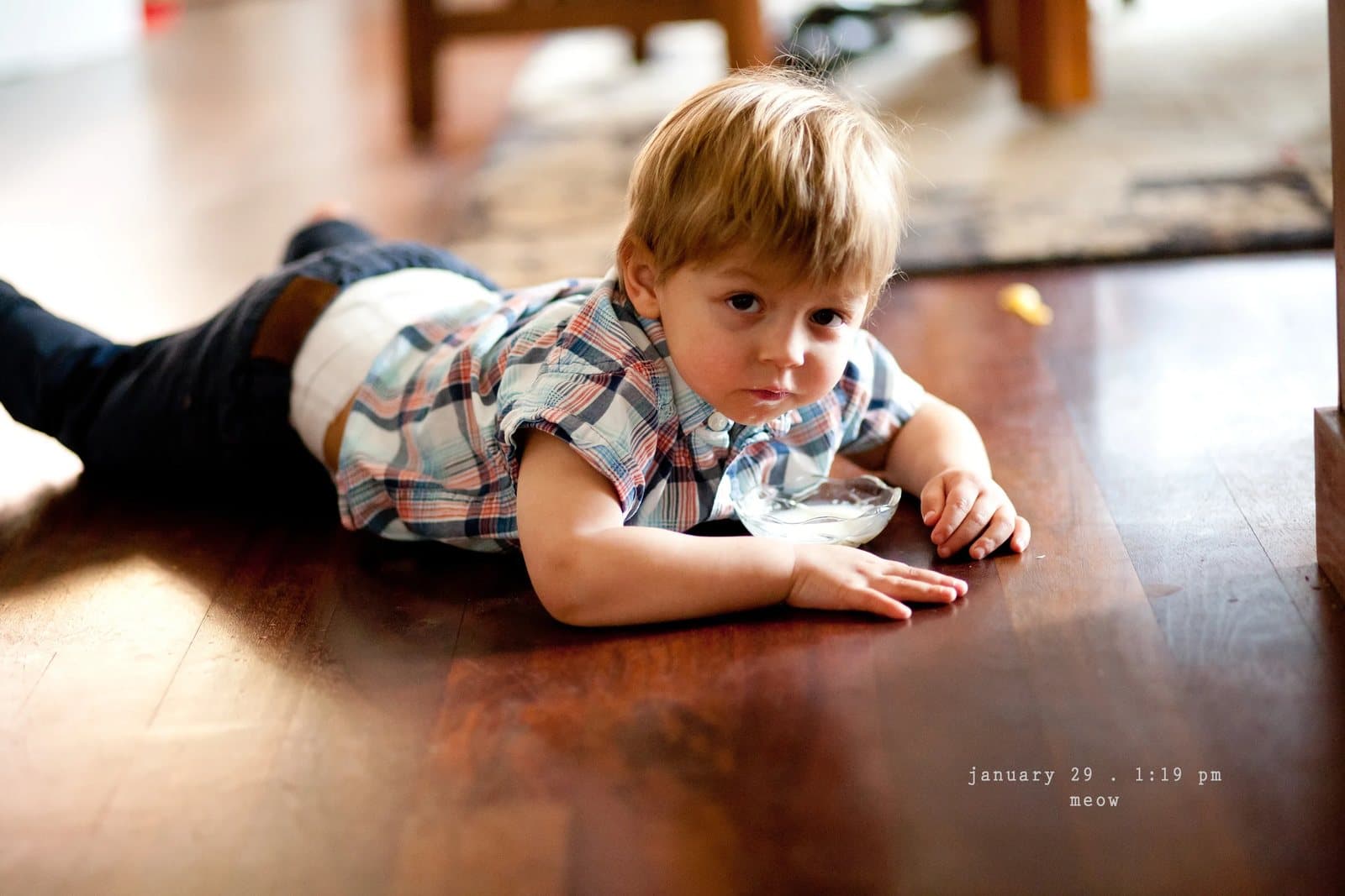 A toddler in a plaid shirt lies on his belly on a wood floor, captioned meow.