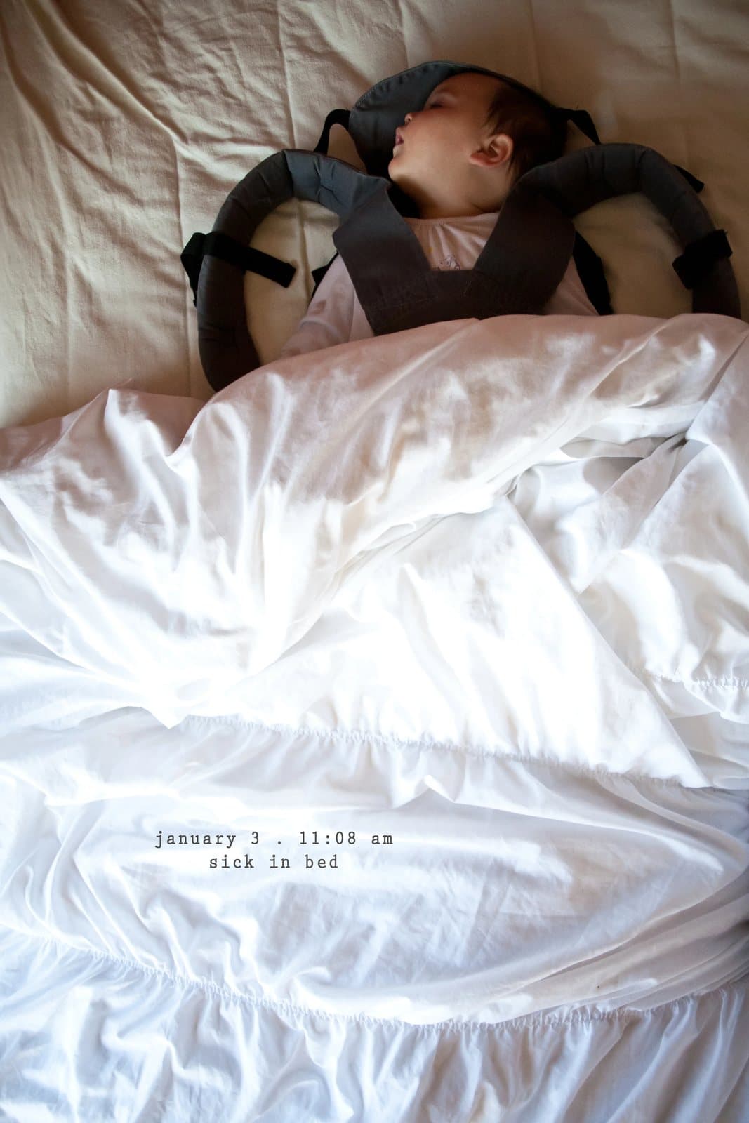 A baby sleeps in a carrier on a bed of white sheets, captioned sick in bed.