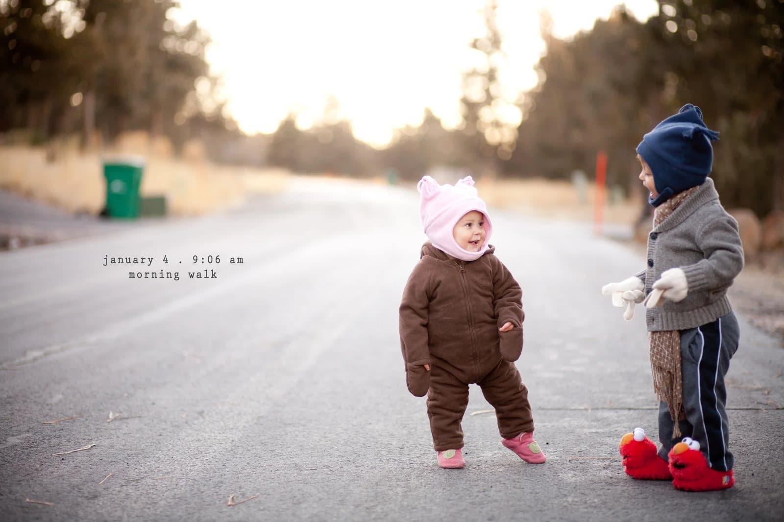 A baby in a pink hat and a toddler in Elmo slippers on a road, captioned morning walk.