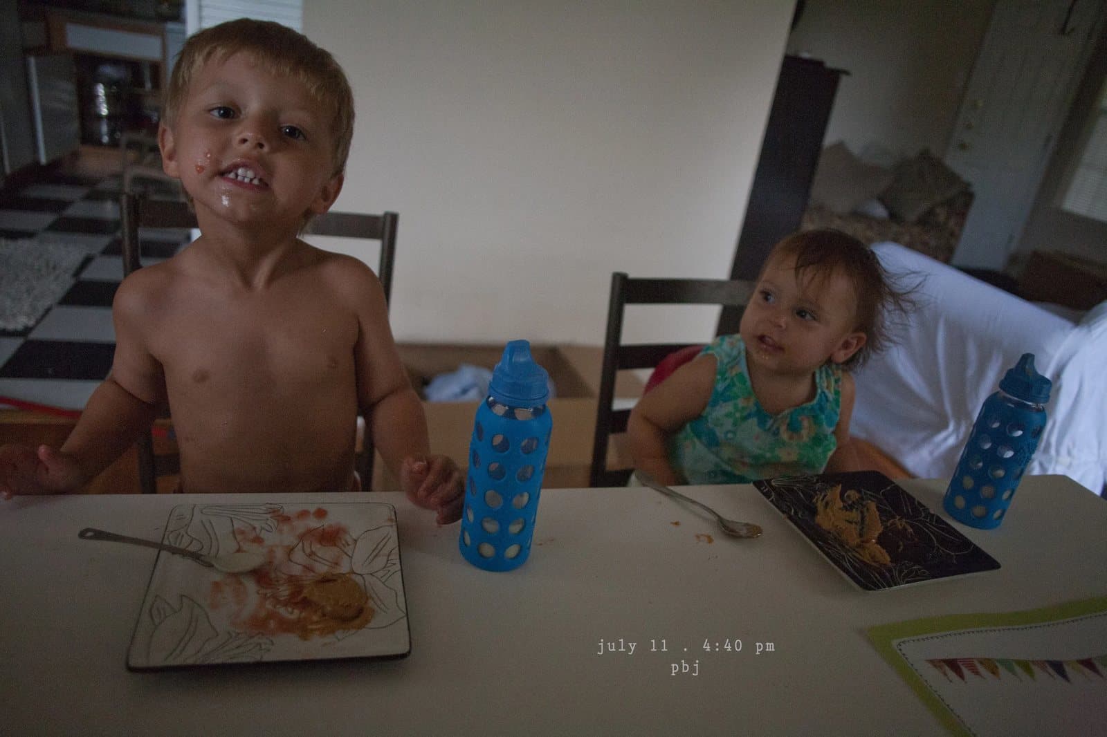 A shirtless boy grins over a messy plate beside a baby at the table, captioned pbj.