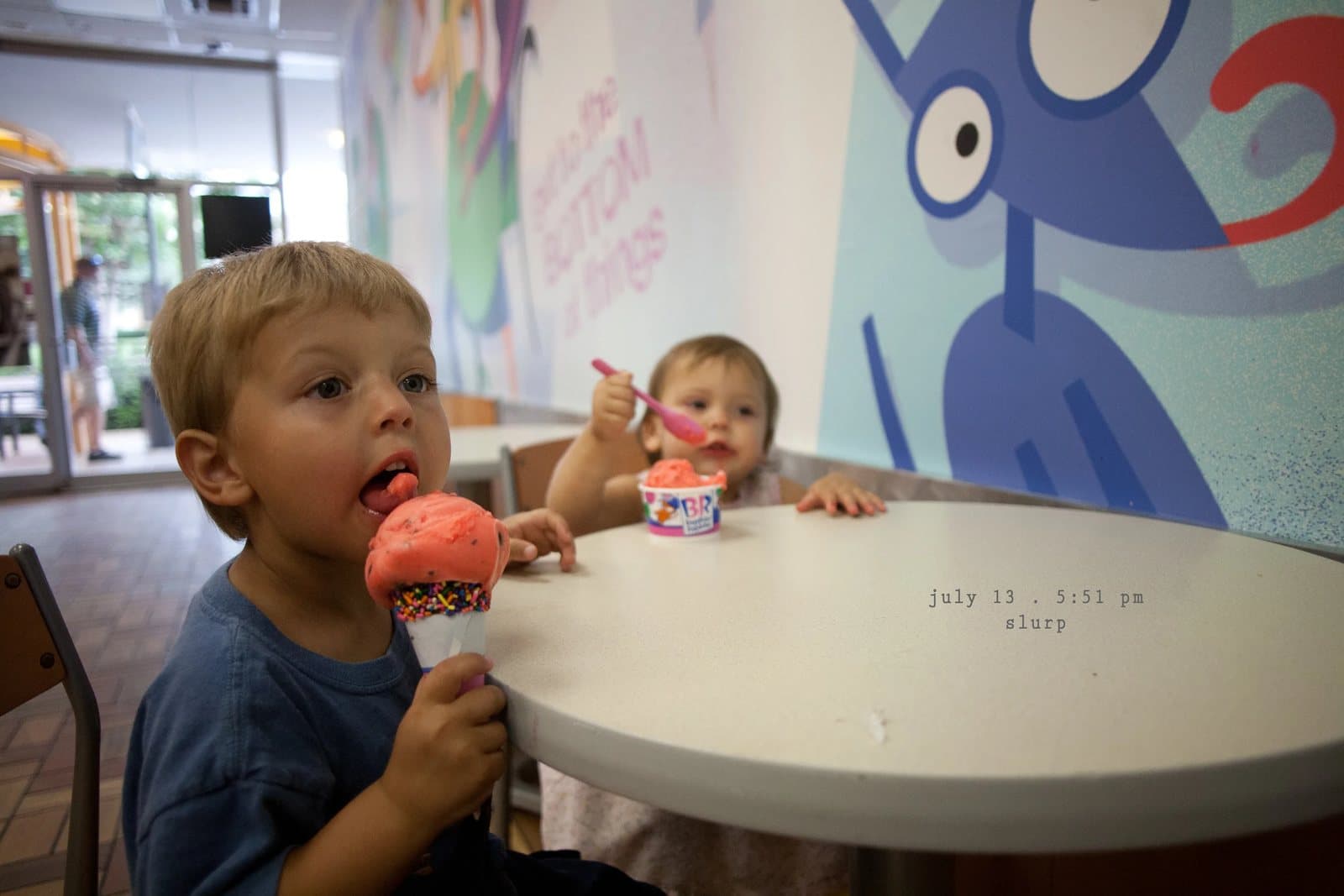 A boy and a baby eat ice cream at a cafe table beside a cartoon mural, captioned slurp.