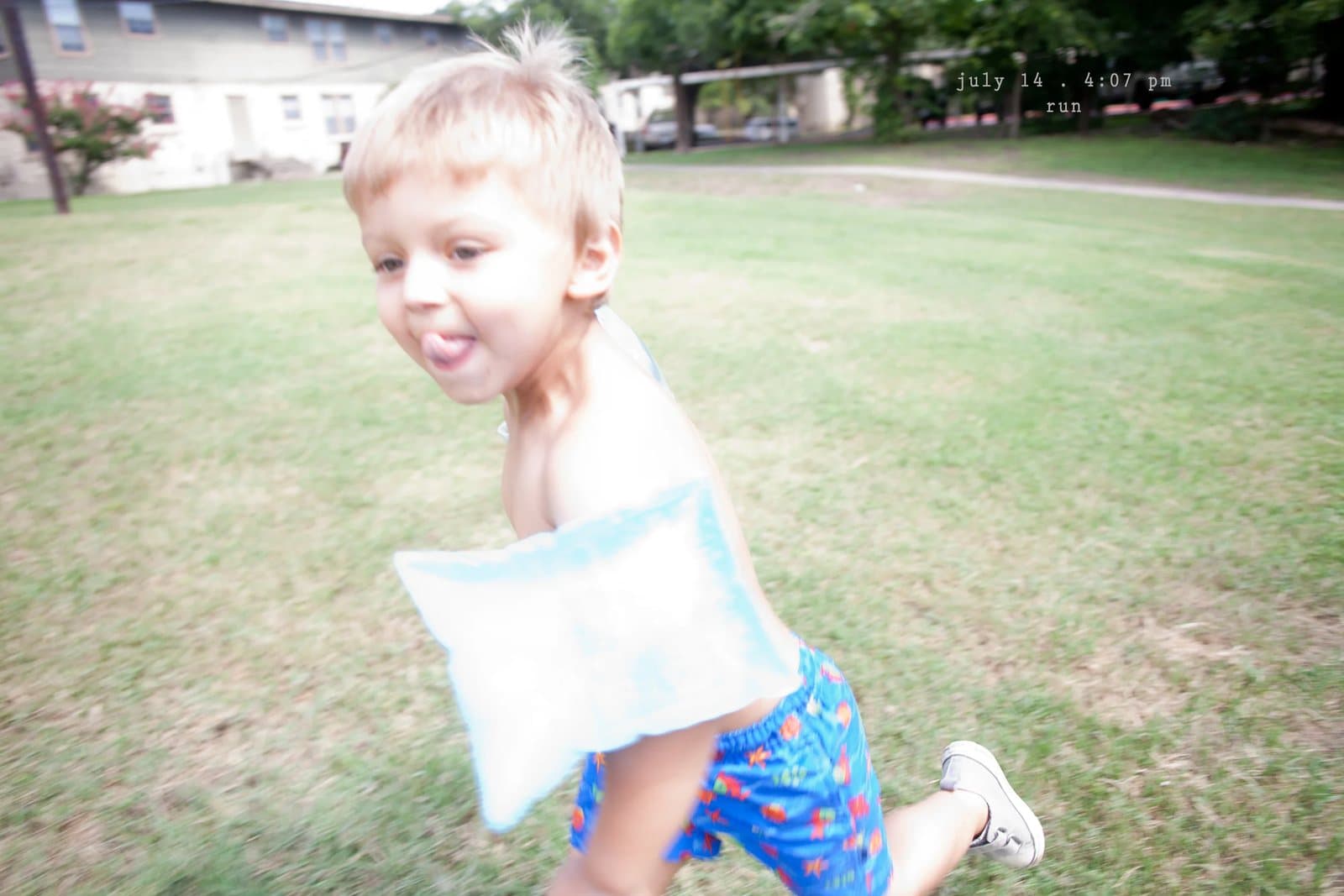 A toddler with a pillow runs across a lawn sticking out his tongue, captioned run.