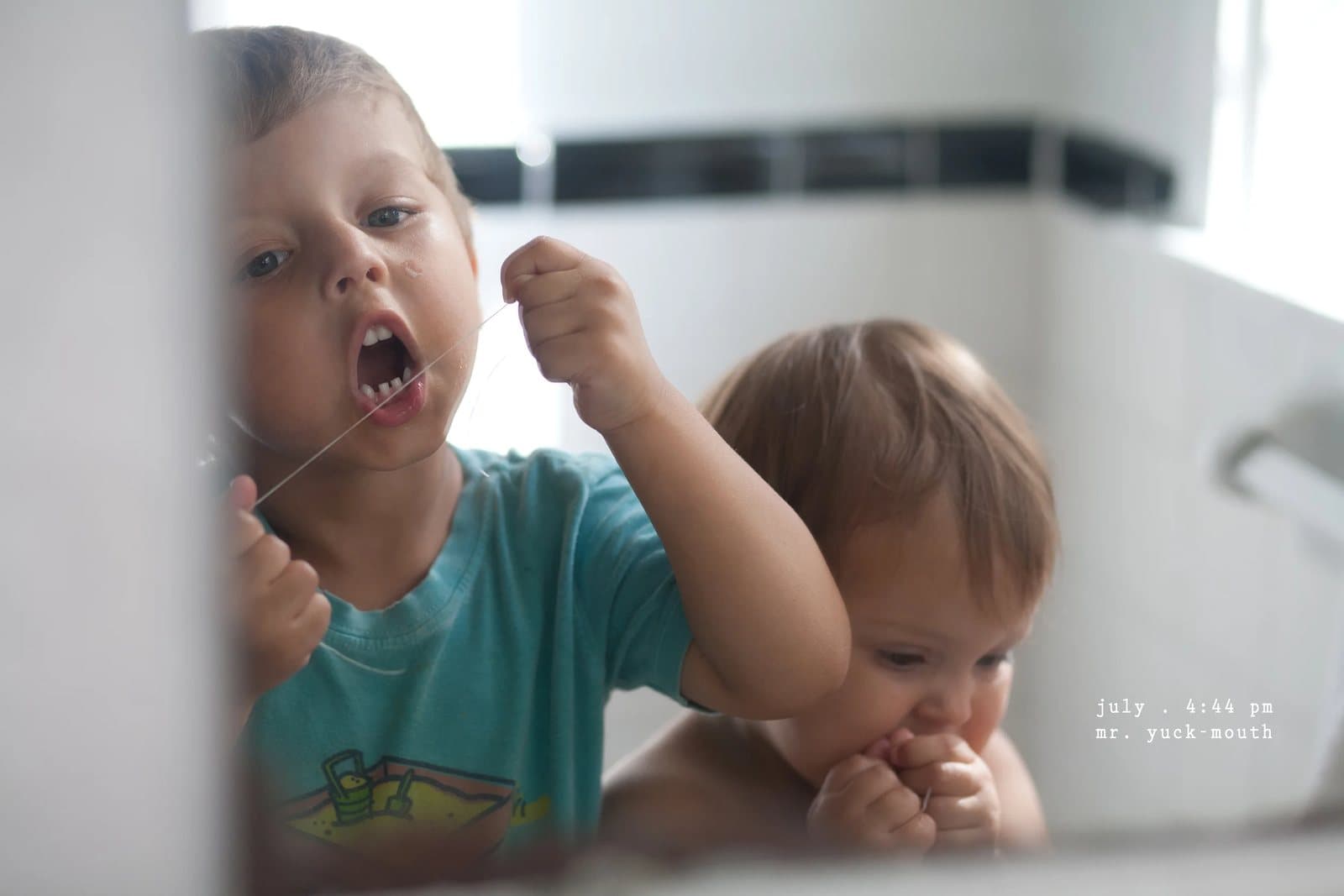 A boy flosses his teeth as a baby leans on his shoulder, captioned mr. yuck mouth.