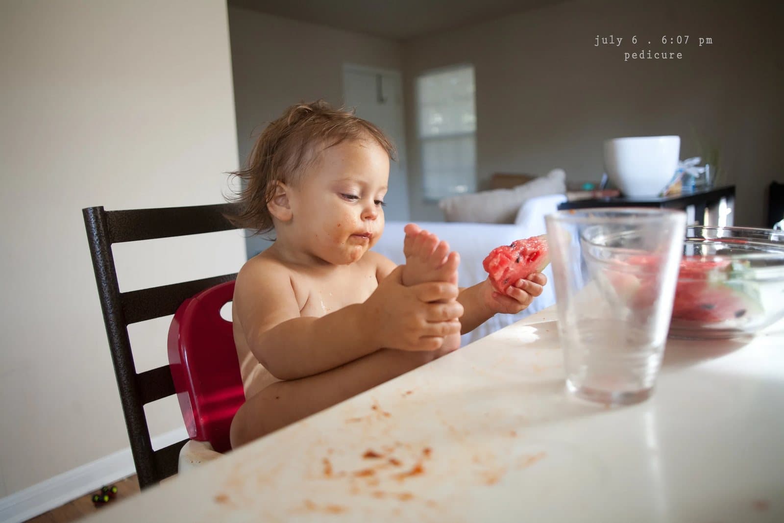 A baby at the table holds her foot and a slice of watermelon, captioned pedicure.