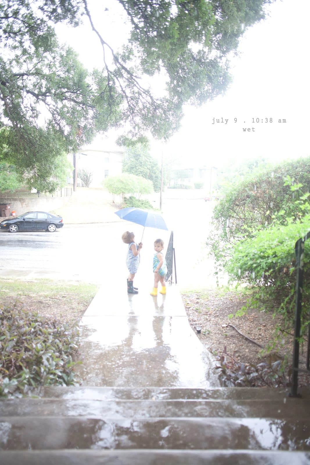 Two small children under a blue umbrella on a rainy sidewalk, captioned wet.
