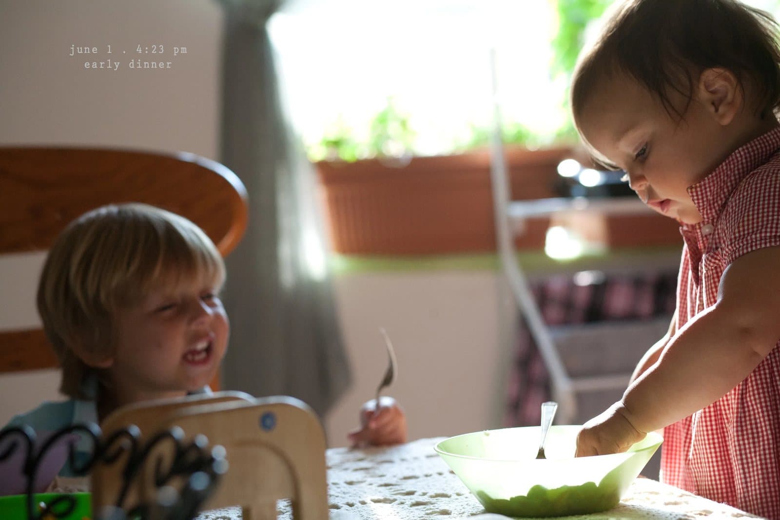 A toddler grins as a baby girl stirs a bowl at the table, captioned early dinner.