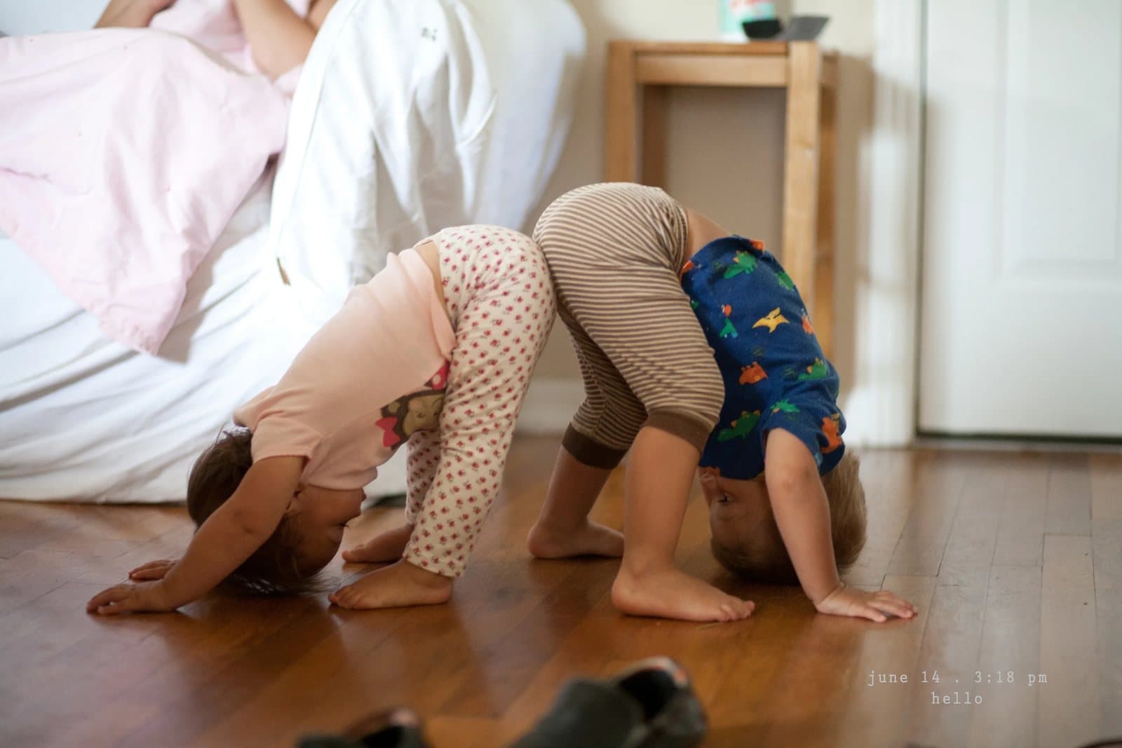 Two toddlers bend over in downward dog poses on a wood floor.
