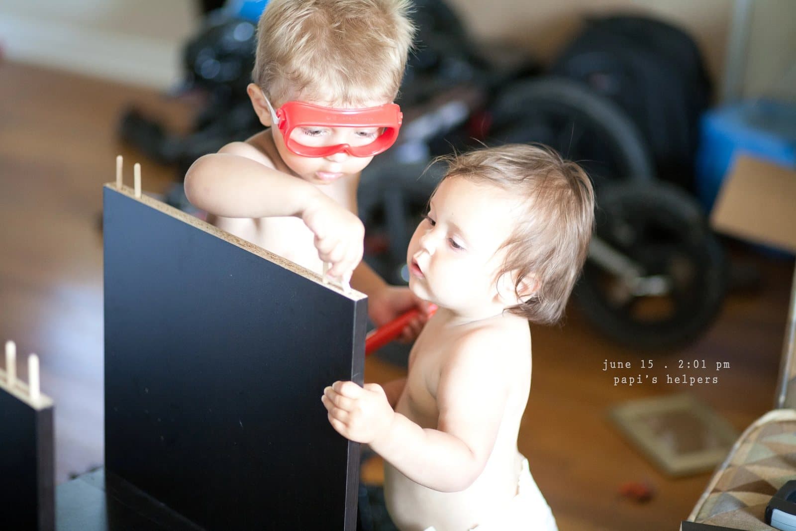 A boy in red goggles and a baby help assemble furniture, captioned papi's helpers.