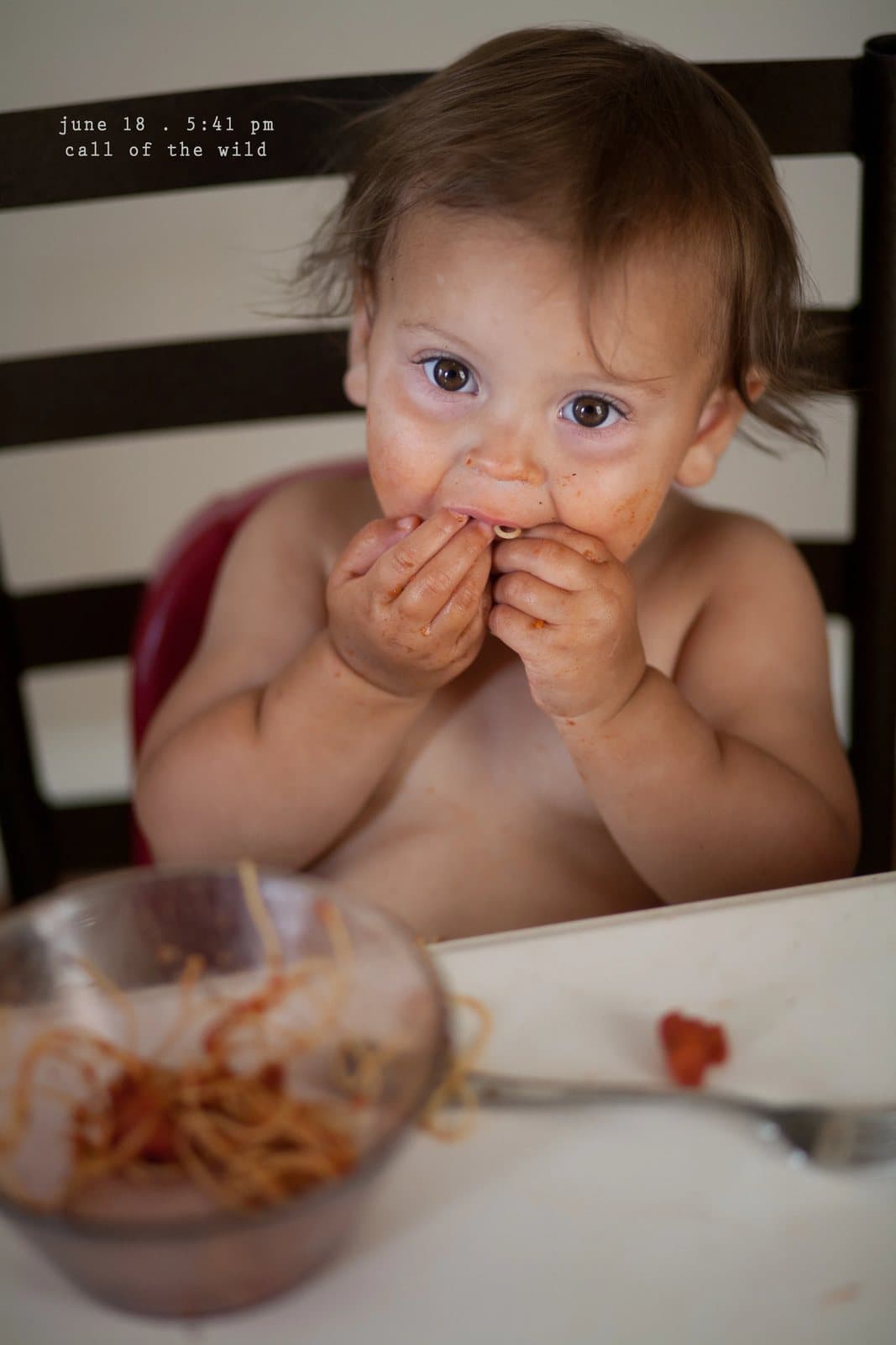 A shirtless baby with spaghetti sauce on her face eats with her hands, captioned call of the wild.