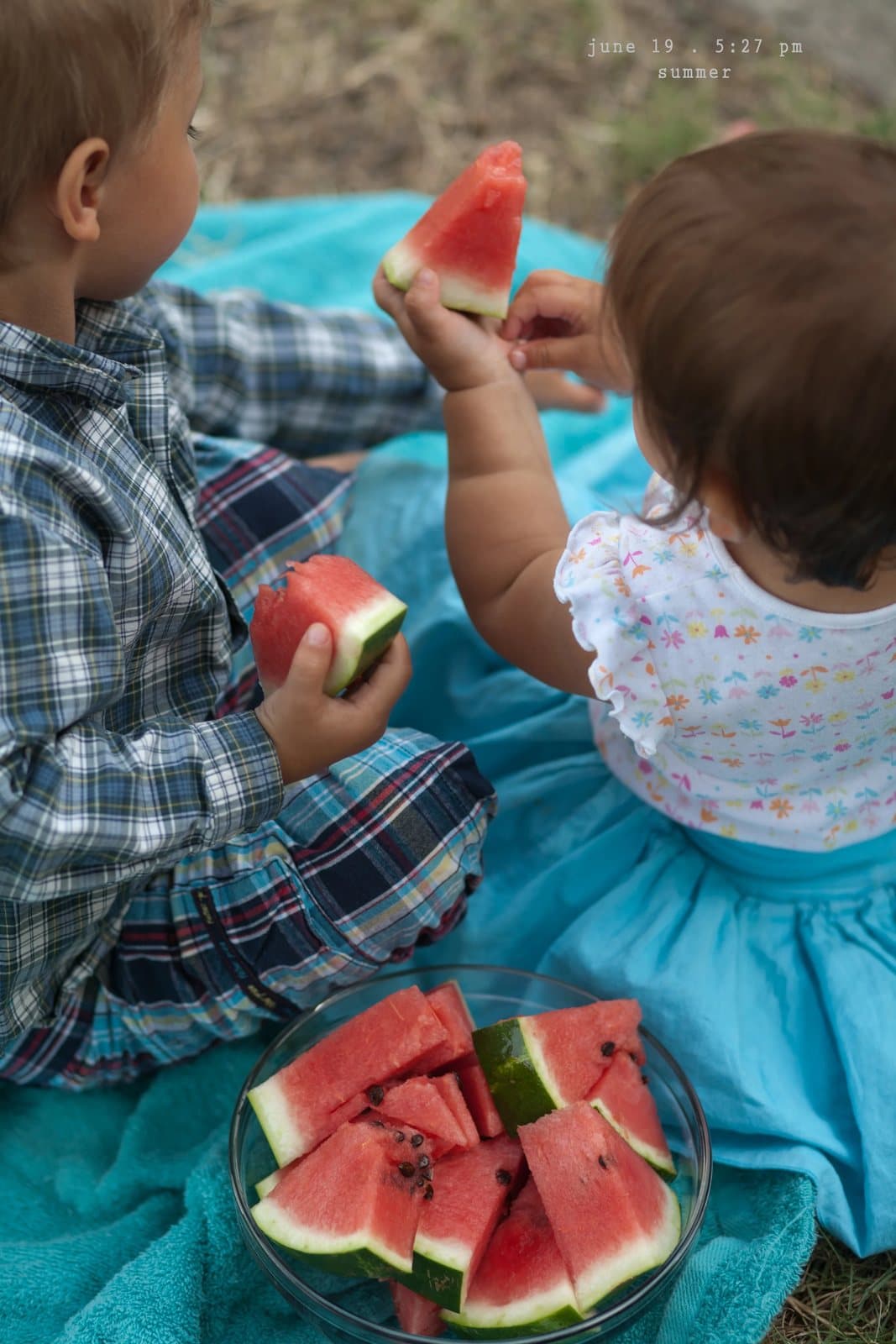 A boy and a baby eat watermelon slices on a blue picnic blanket, captioned summer.