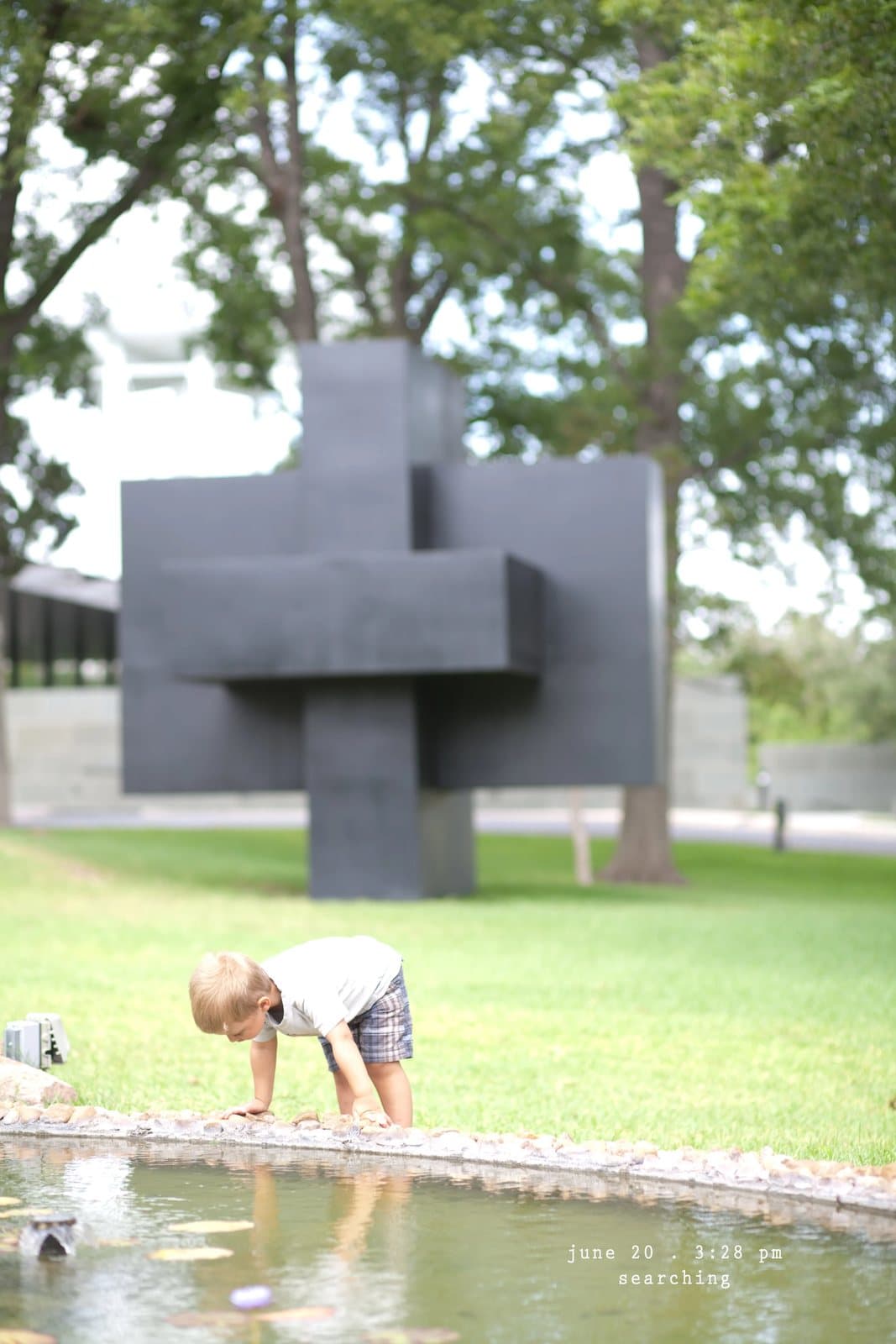 A toddler crouches by a pond near a large dark geometric sculpture, captioned searching.
