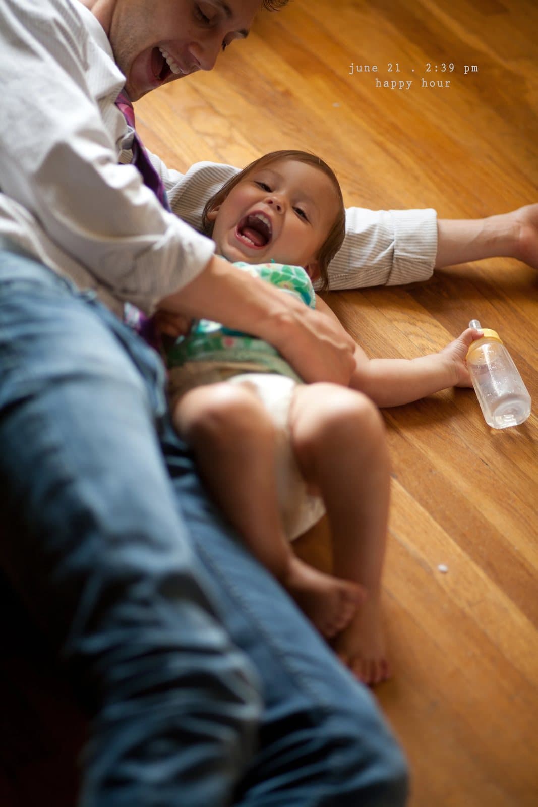 A man tickles a laughing baby on a wood floor beside a bottle, captioned happy hour.