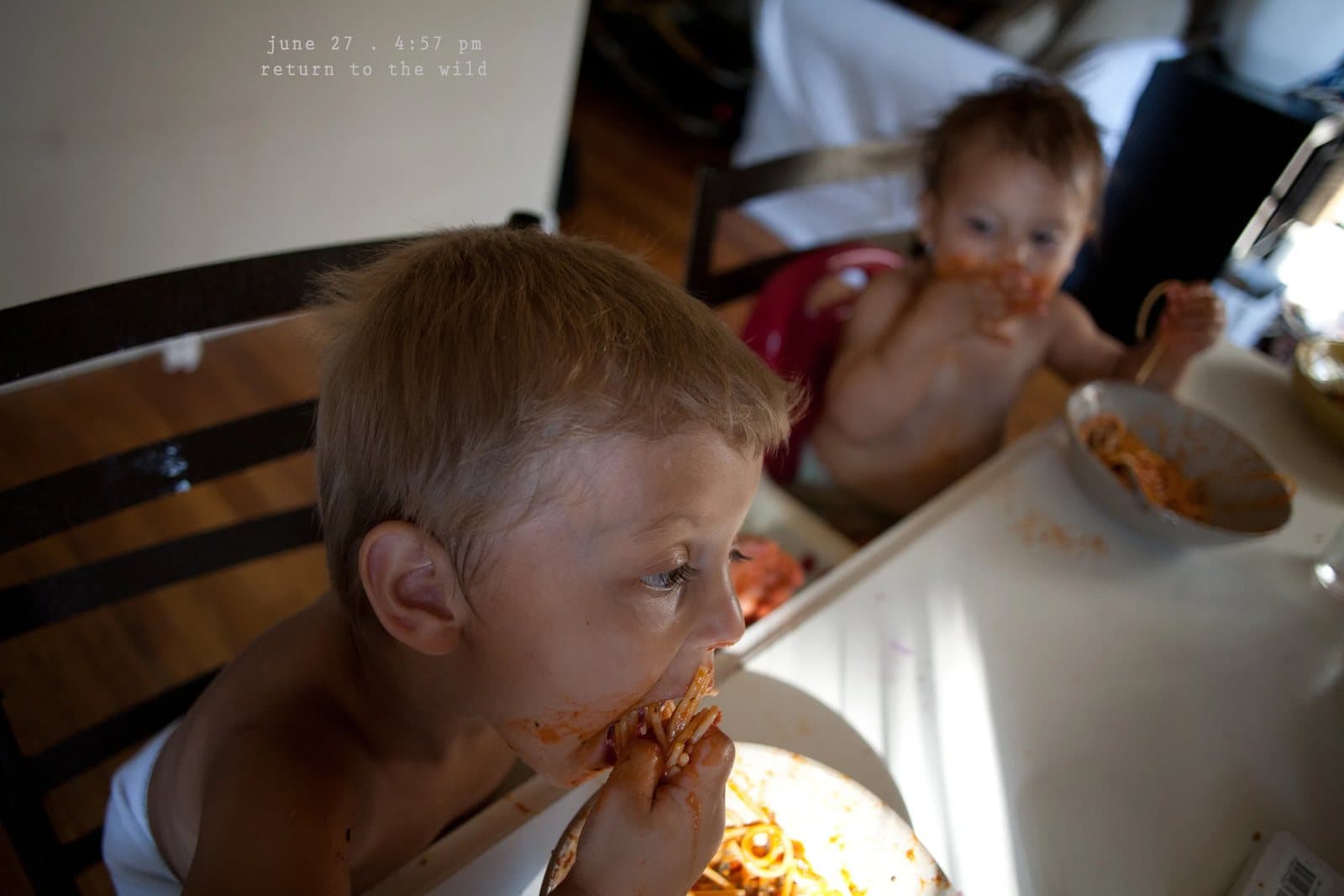 A boy and a baby eat spaghetti messily at the table, captioned return to the wild.