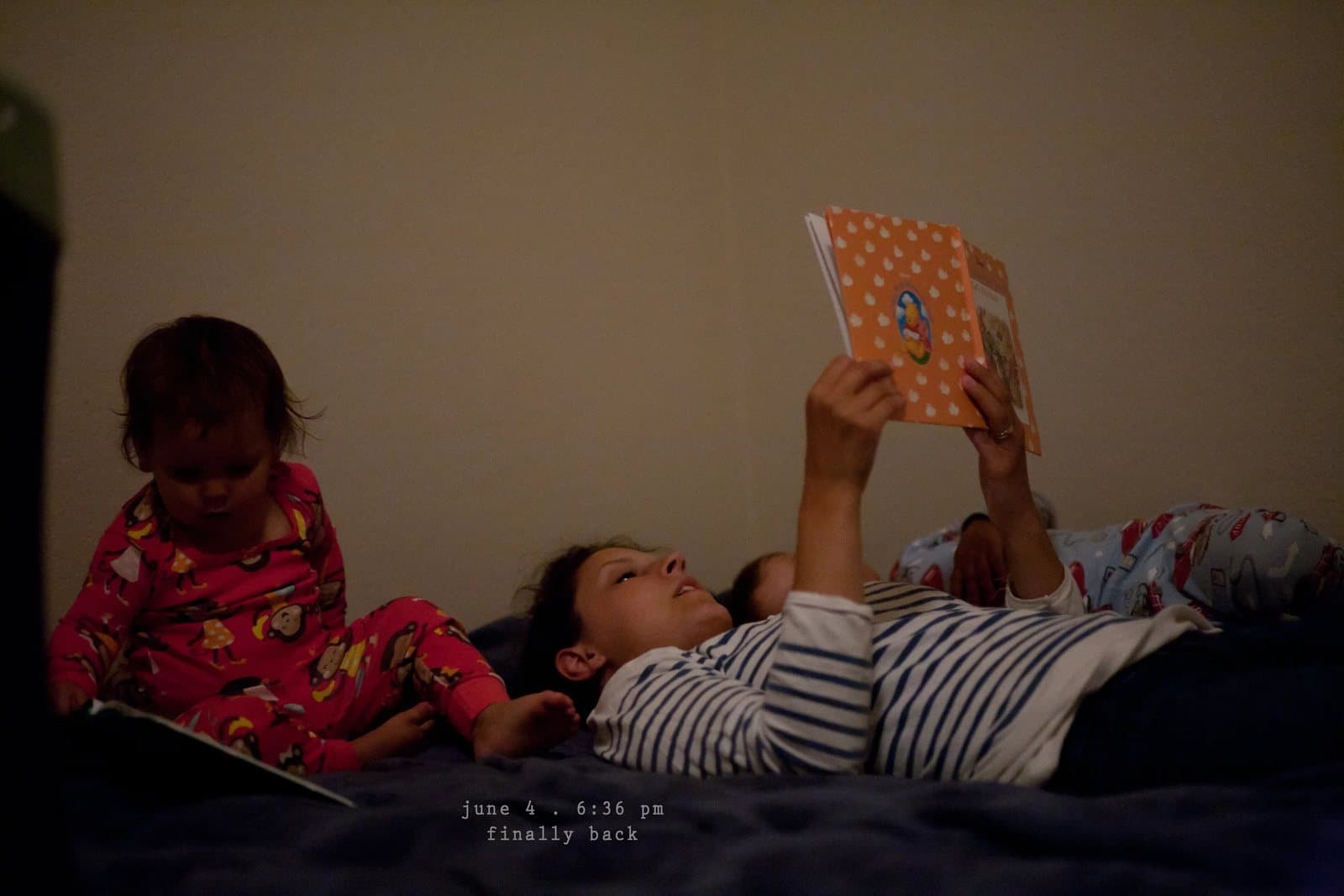 A woman reads a picture book in bed beside two children in pajamas, captioned finally back.