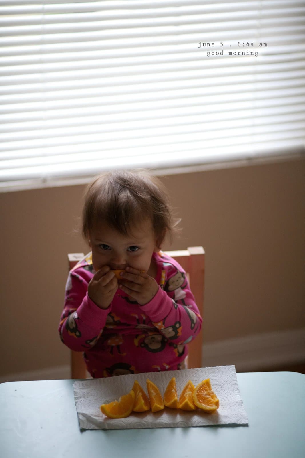 A baby in pink pajamas eats orange slices at a table by a window, captioned good morning.