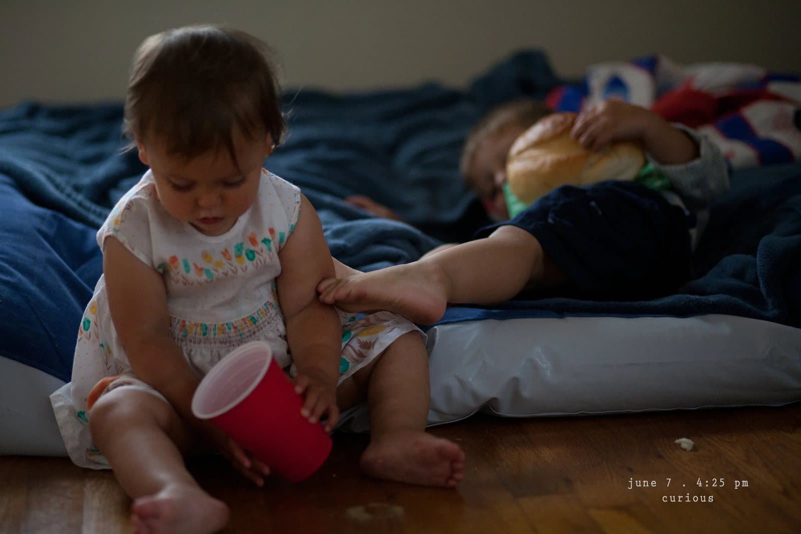 A baby in a sundress plays with a red cup beside a sleeping child, captioned curious.
