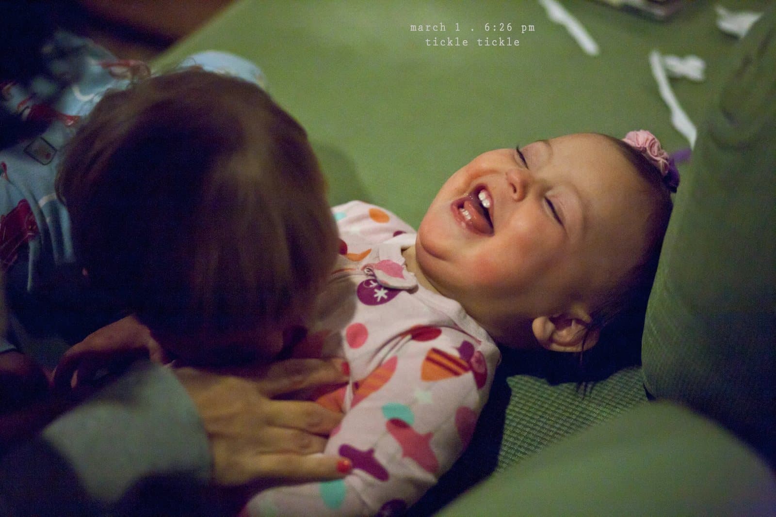 A laughing baby in pajamas is tickled on a green couch, captioned tickle tickle.