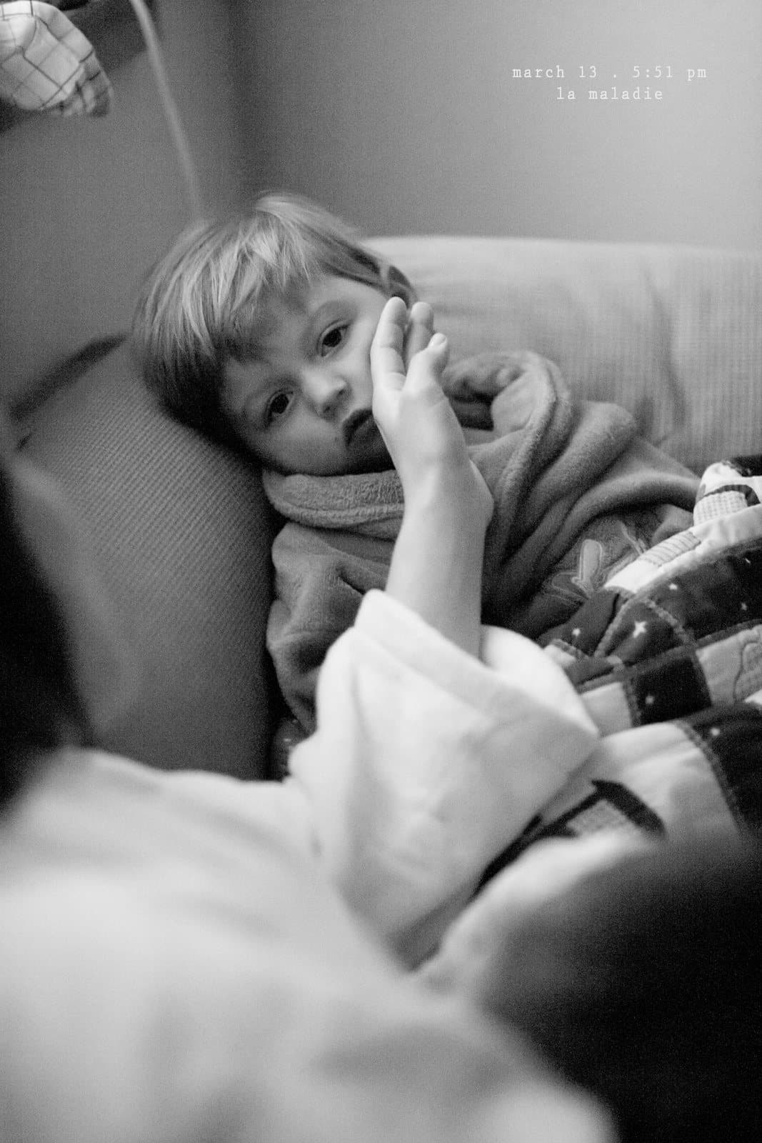 A sick toddler lies on a couch under a blanket in black and white, captioned la maladie.