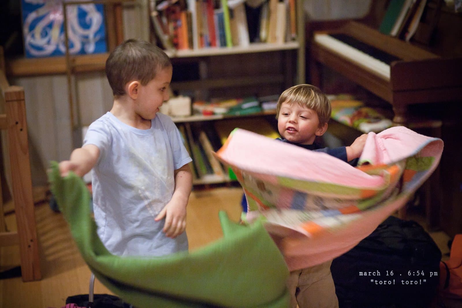 Two boys swing colorful blankets like capes in a playroom, captioned toro! toro!