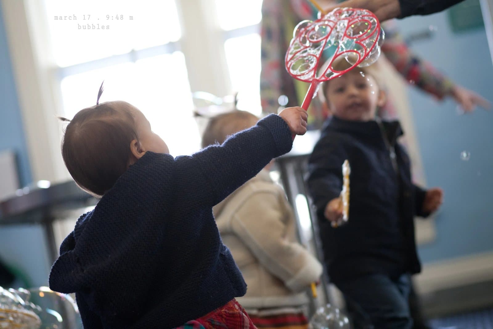 Children reach for soap bubbles from a wand in a bright room, captioned bubbles.