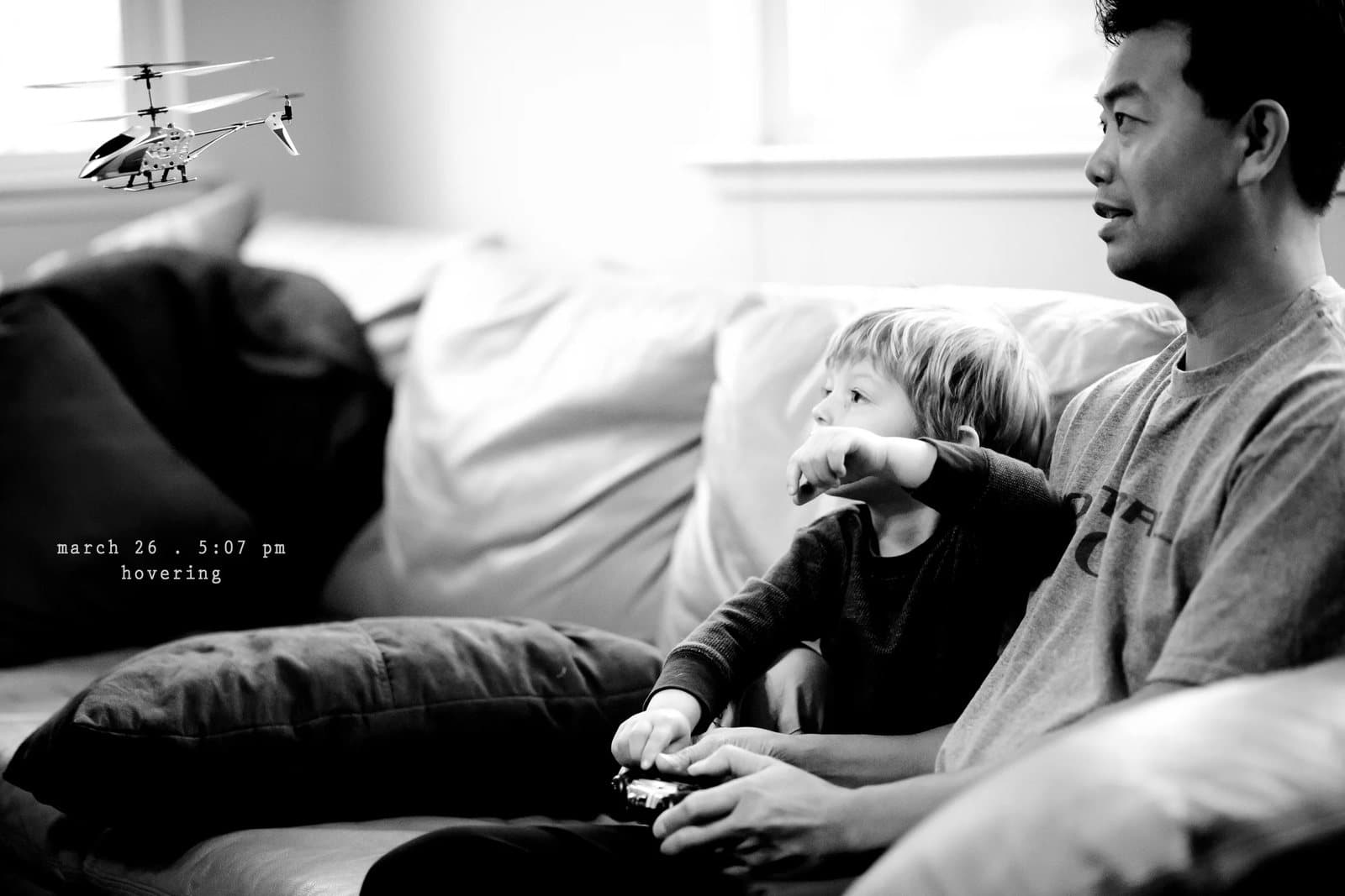 A toddler and a man on a couch watch a toy helicopter fly, captioned hovering.